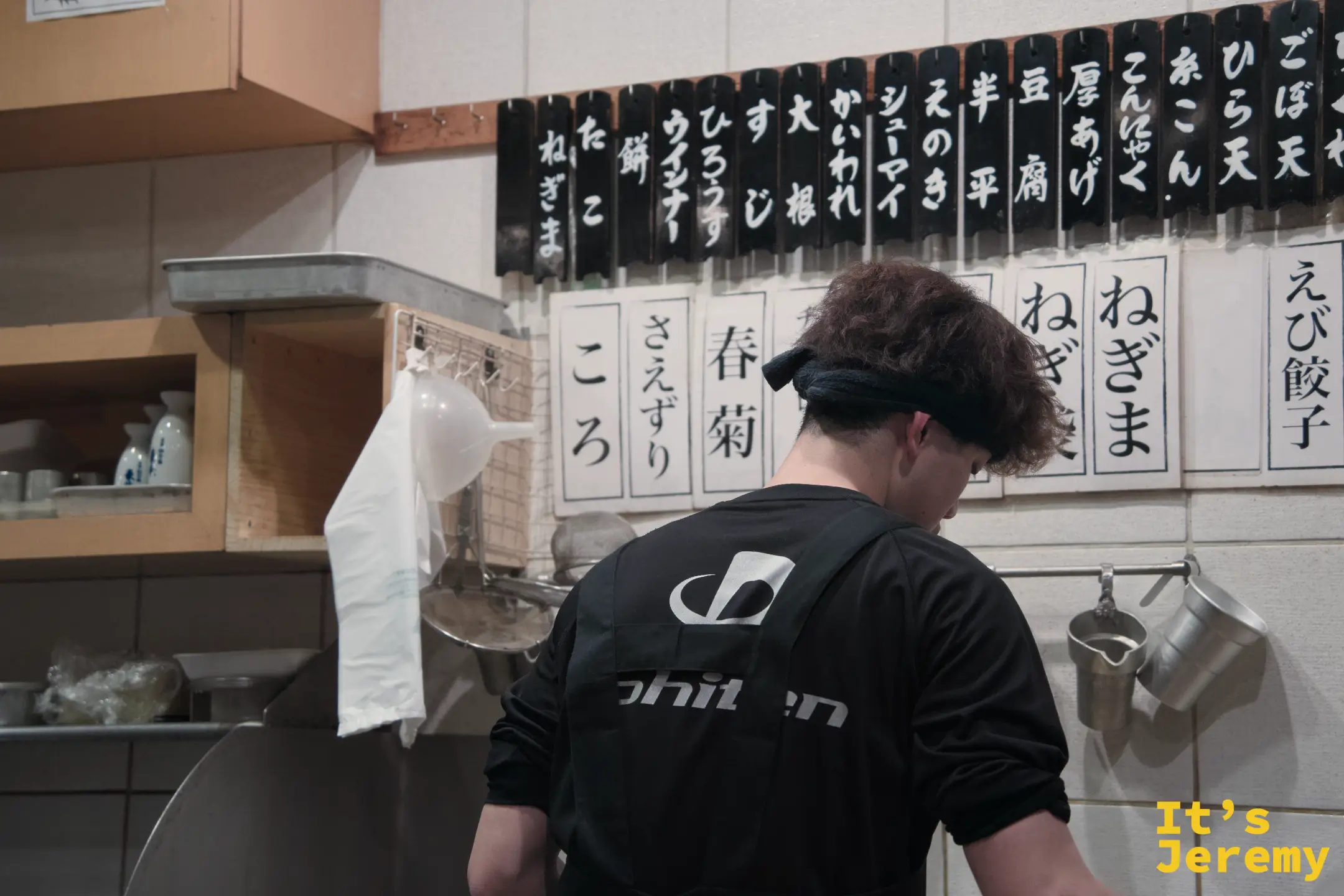 師傅背對鏡頭作業，身後牆上貼著印刷字卡，列出所有おでん品項