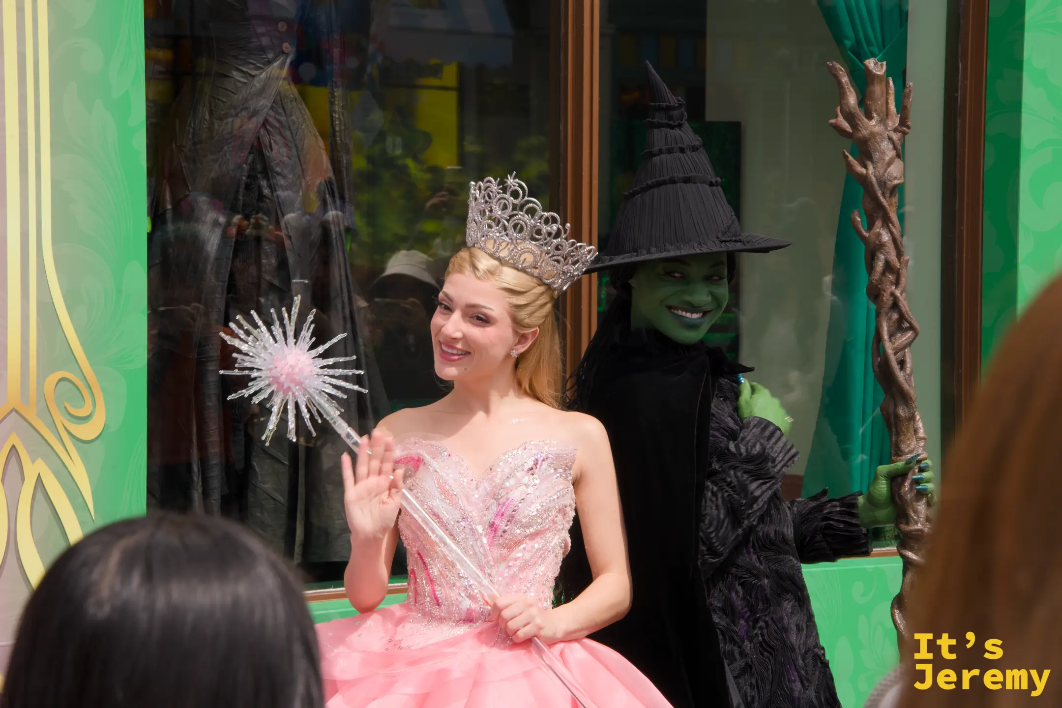 Glinda 與 Elphaba 並肩而立，一個穿著粉白蓬裙手持水晶魔法棒，另一個穿著黑色斗篷舉著老樹杖，形成強烈對比
