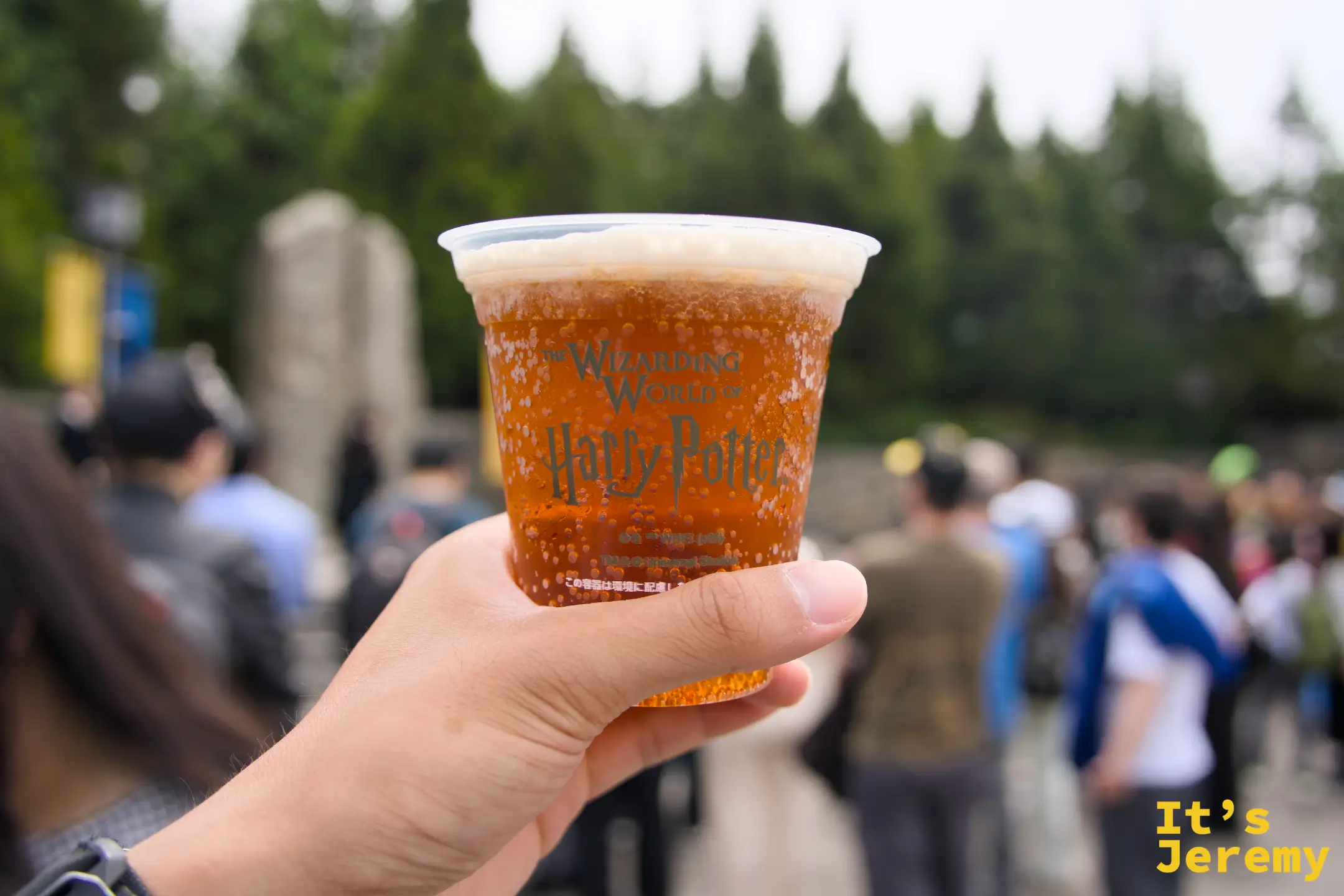 奶油啤酒，杯身印著「The Wizarding World of Harry Potter」字樣，橘紅色液體配上白色奶泡，在霍格莫德村的石牆前舉起來拍就很好看
