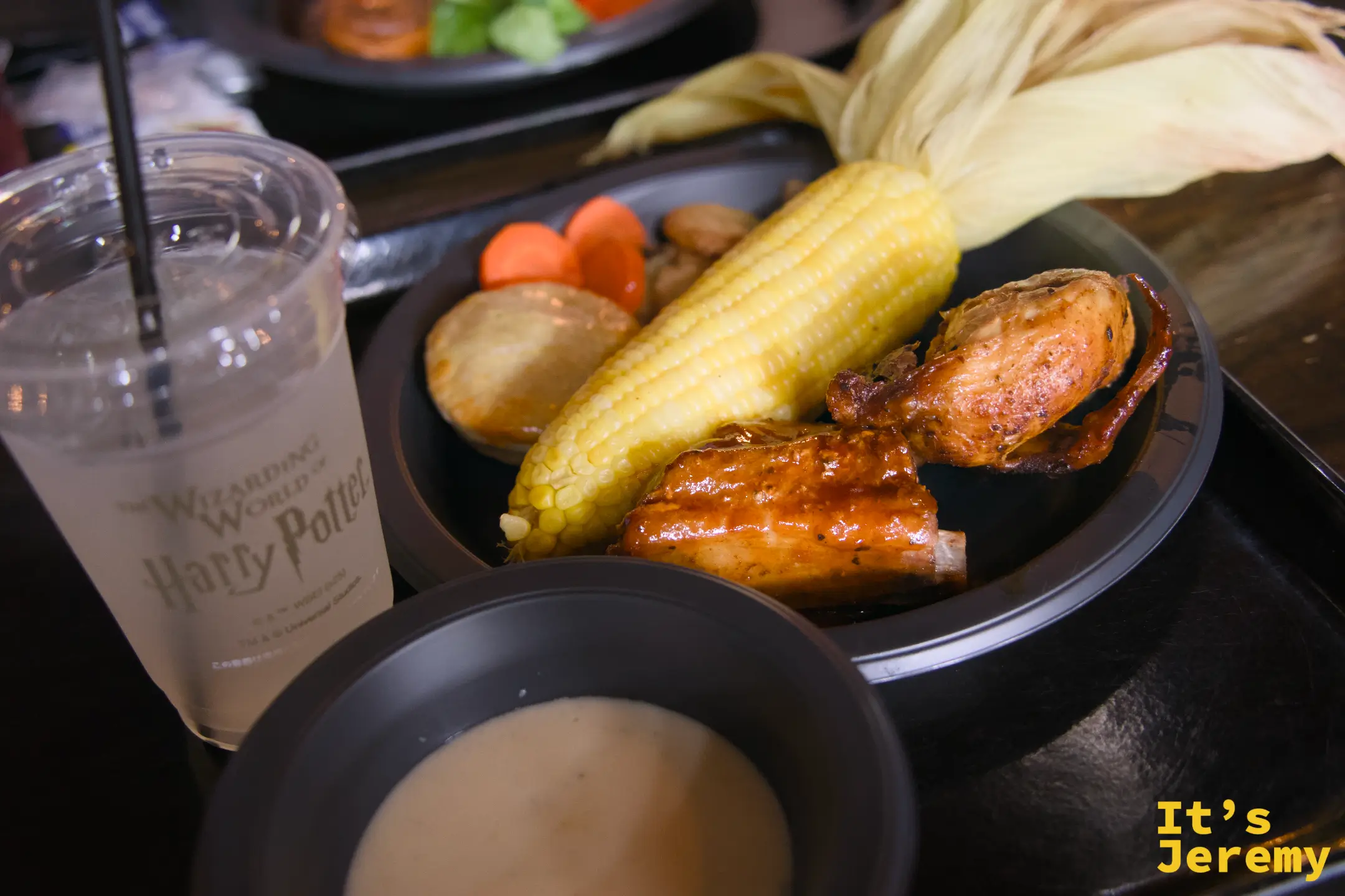 三本の箒™ 烤雞與豬肋排套餐，黑色餐盤盛著烤雞塊、豬肋排、玉米、馬鈴薯，旁邊是 Harry Potter 主題的冰杯