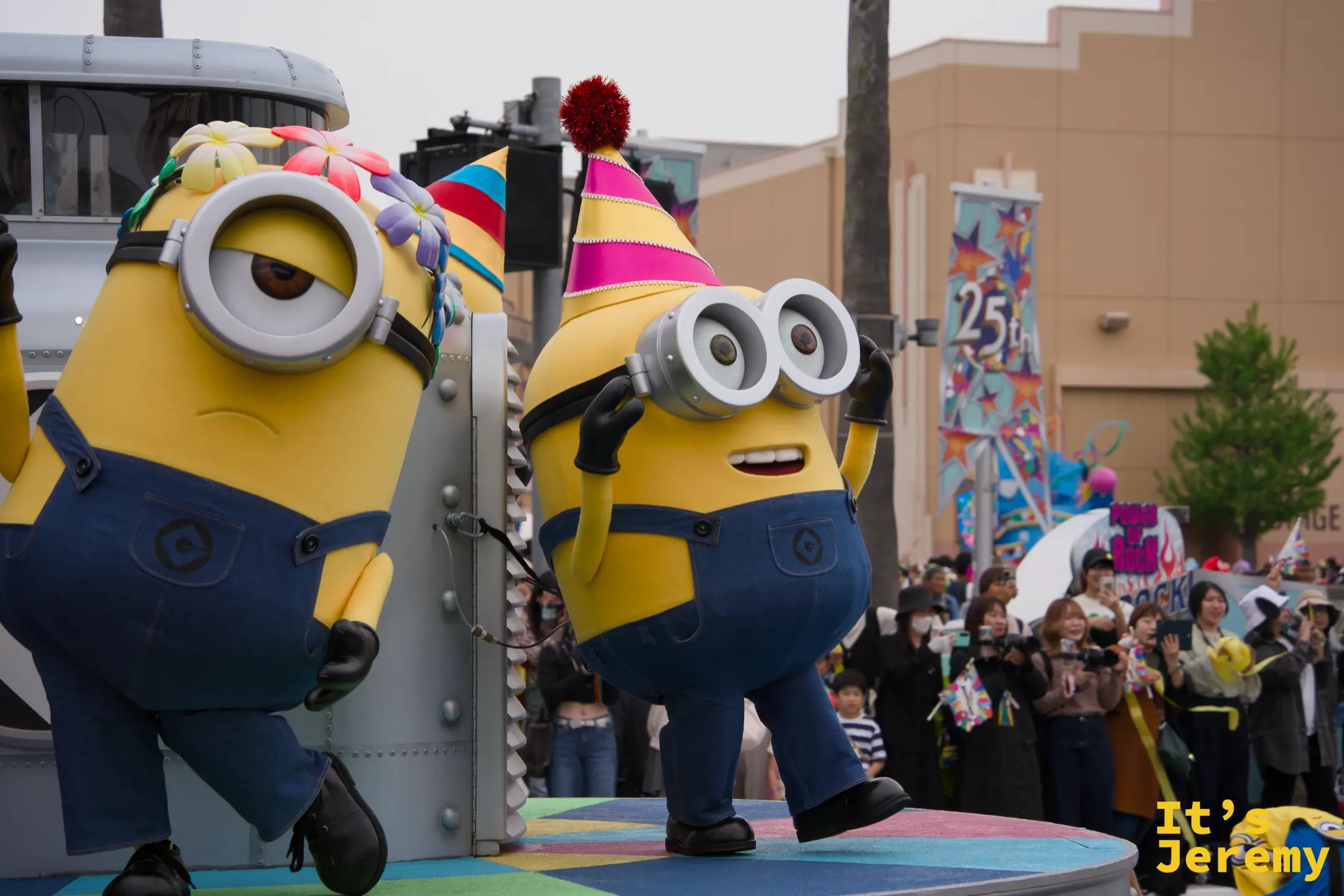 一隻 Minion 從花車旁的路燈基座探出身來，驚訝表情對著觀眾，25th 裝飾旗幟在背後飄揚
