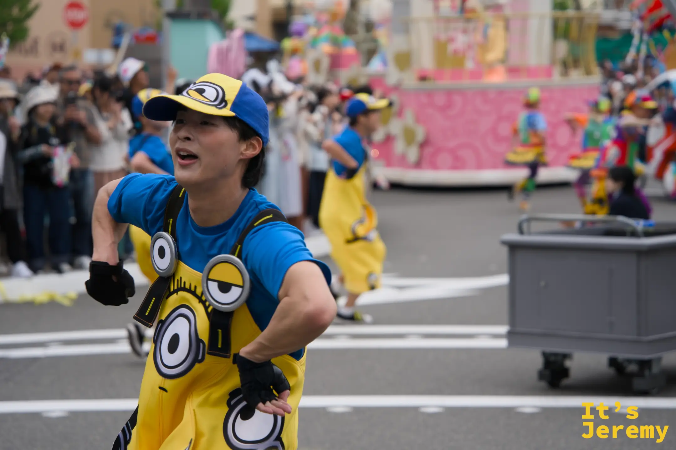 一位身穿 Minion 服裝的舞者在街道上奔跑，後方是色彩繽紛的遊行花車