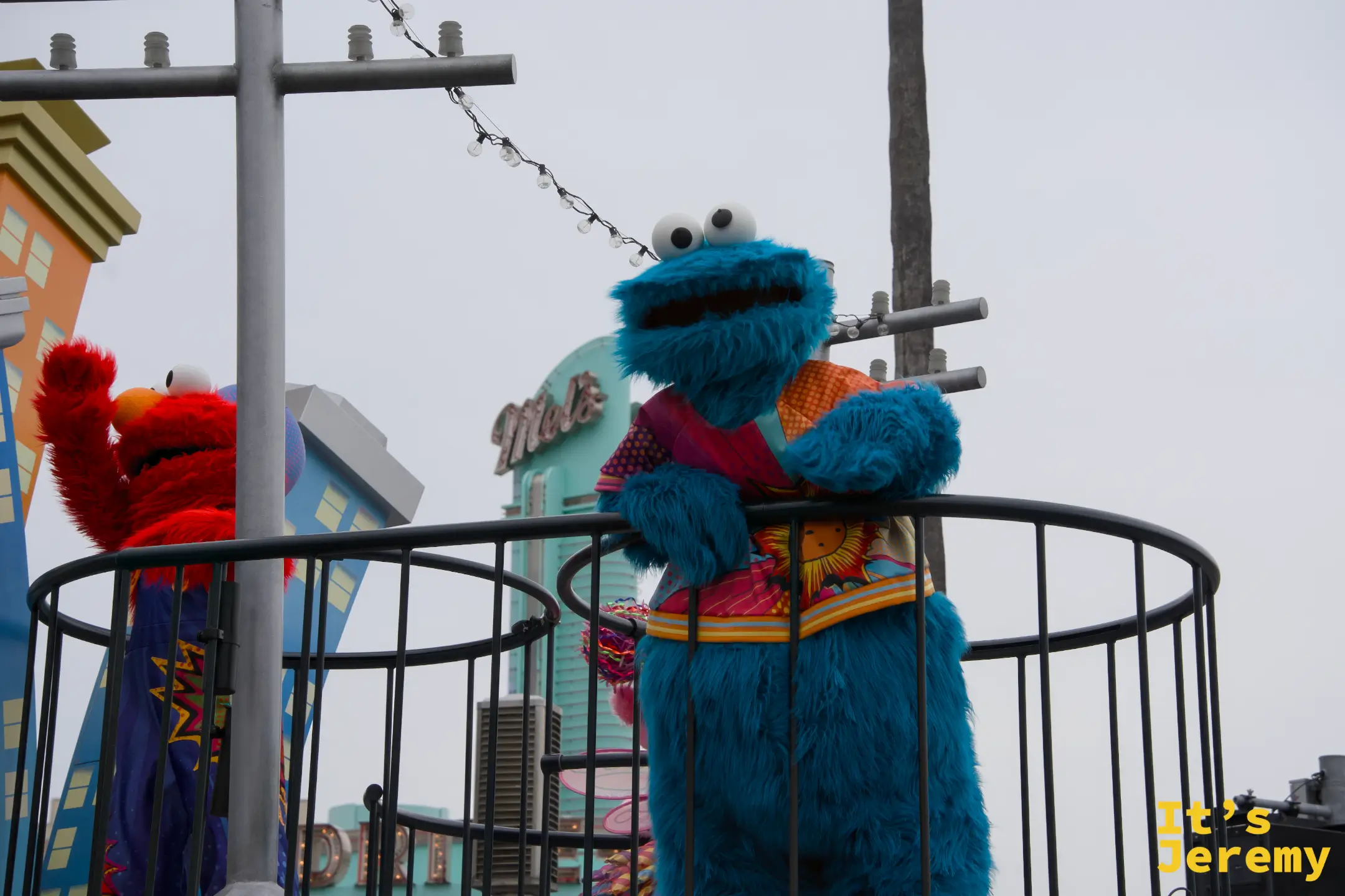 Cookie Monster 與 Elmo 並肩站在花車上，背景是芝麻街特色的彩色磚牆建築，遠處是 Mel
