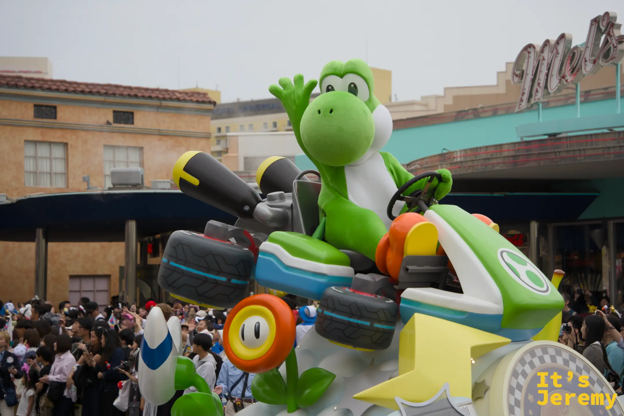 Yoshi 特寫，綠色身軀坐在 Mario Kart 賽車裡，對著觀眾揮手，Mel