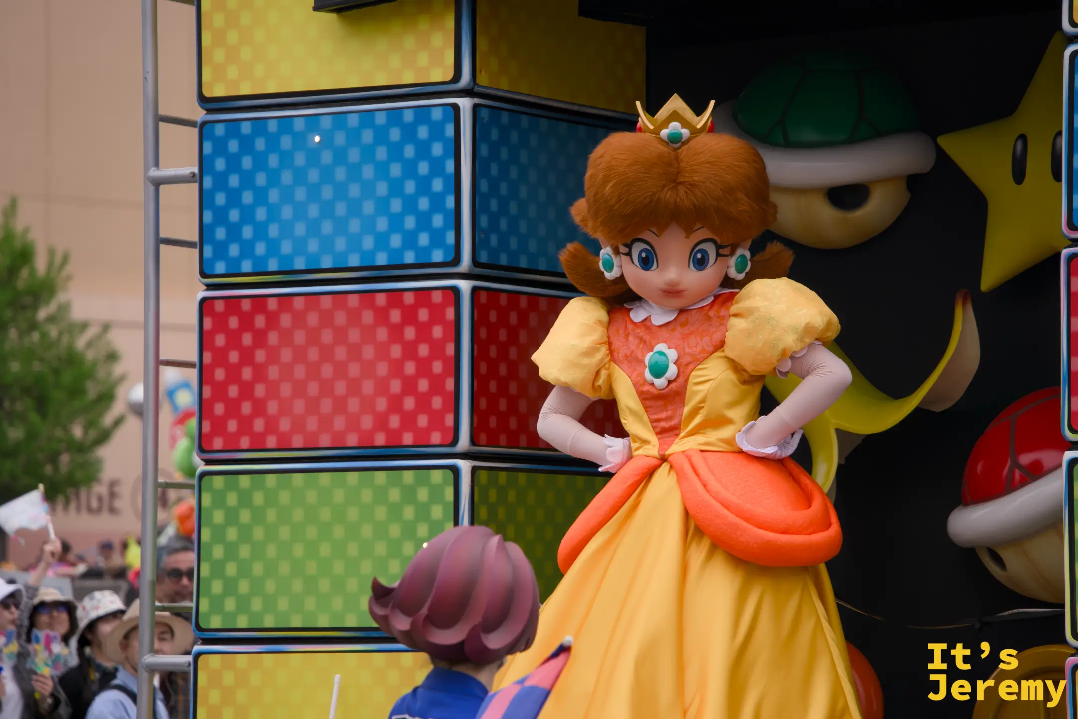Princess Daisy 單獨特寫，橘黃蓬裙配上 D 字項鍊，站在馬賽克磚塊花車邊沿，角落還隱約可見一顆 Poké Ball