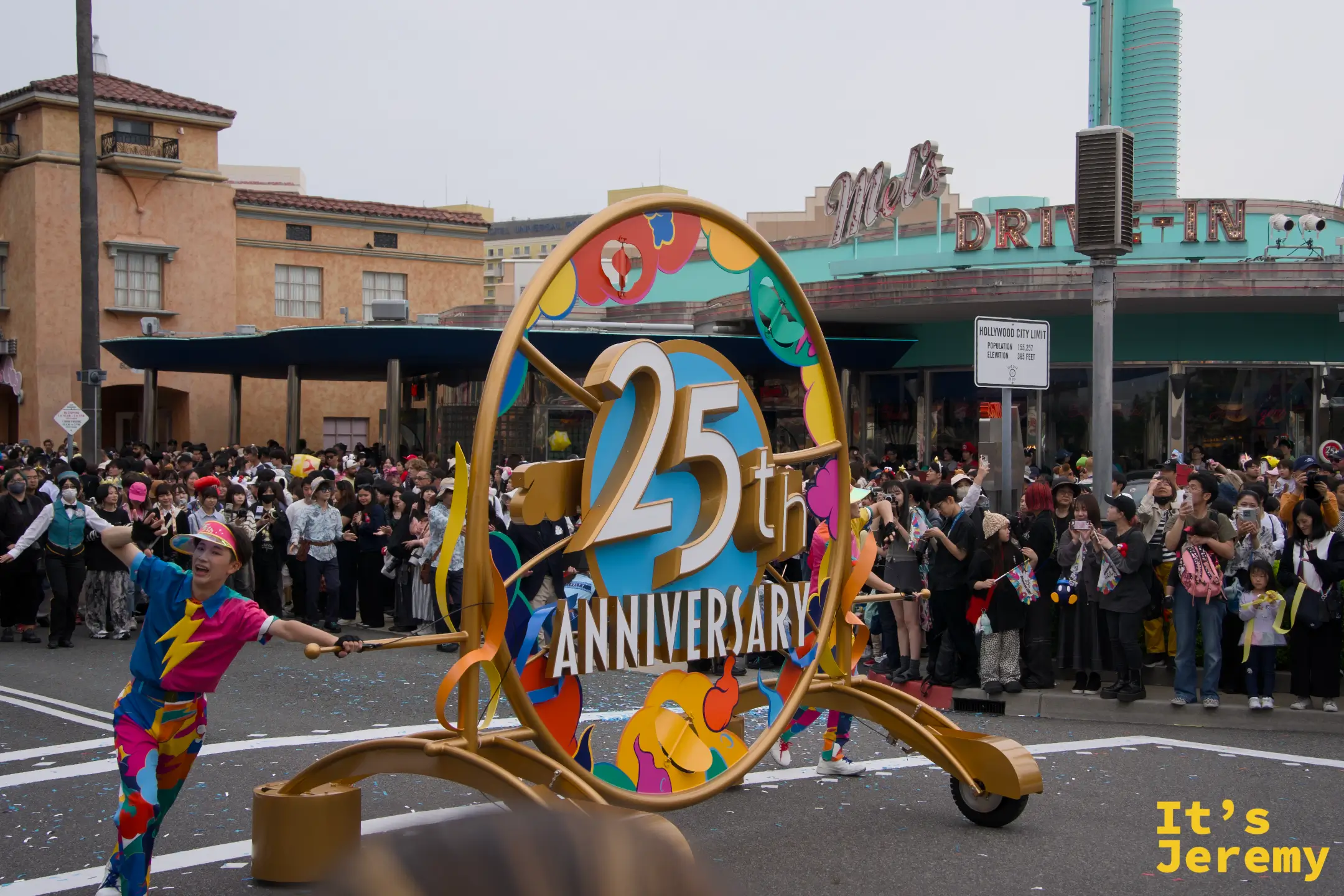 25th Anniversary 金色圓形標誌特寫，演員在兩側舉著標誌在街道上走動