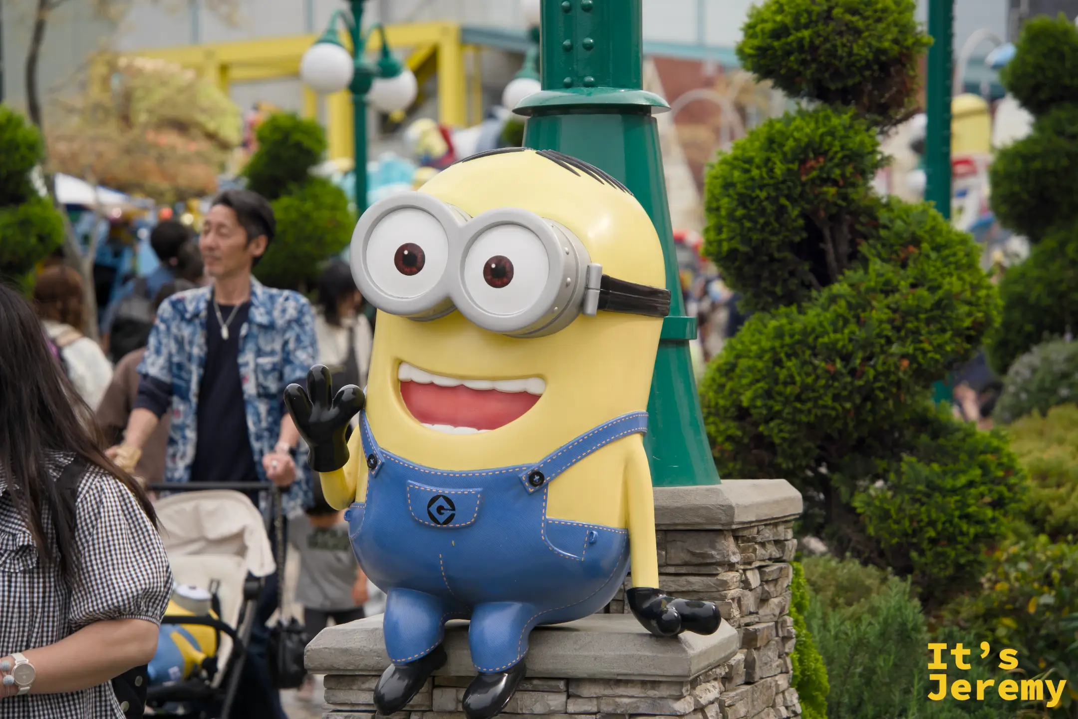 Minion 雕像坐在庭院的低矮石墩上，背後是黃綠相間的 Minion Park 景觀