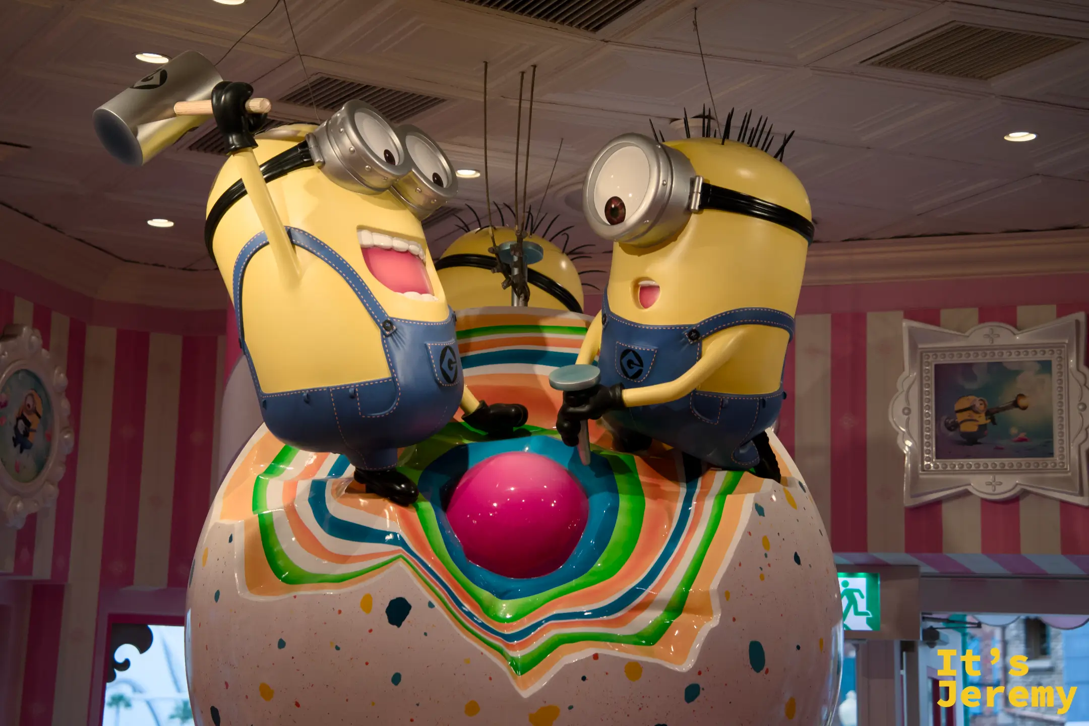 三隻 Minion 雕像攀在甜甜圈球體上，表情各異，從天花板俯瞰整個粉色系糕點風格店內