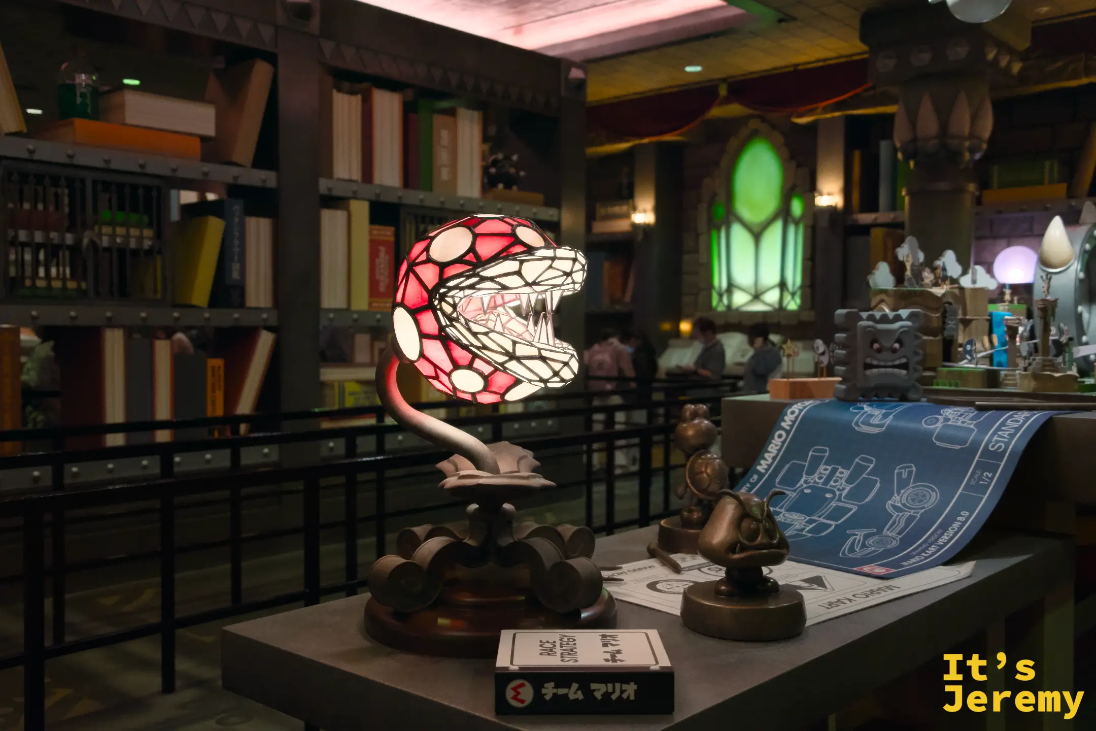 SNW 商店內的 Piranha Plant（吃人花）彩繪玻璃台燈，旁邊散落著 Mario Motocross 設計圖，整個區域的道具設計充滿細節