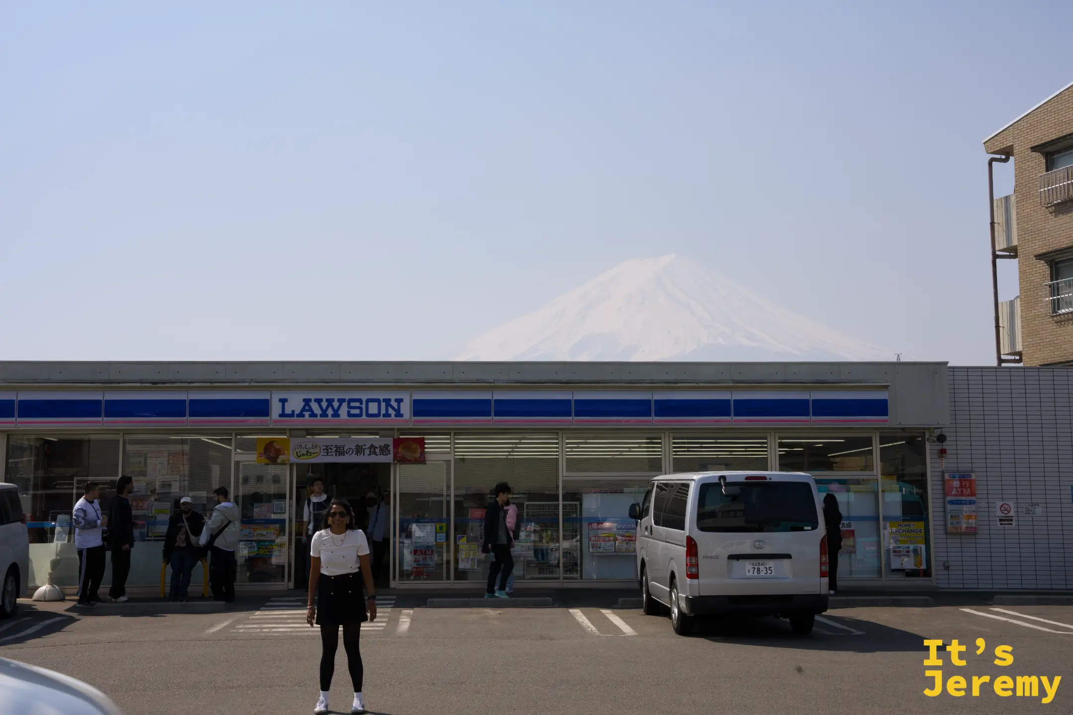 Lawson 便利商店，屋頂後方的富士山被沙塵壓成淡灰色輪廓，停車場有車輛與行人