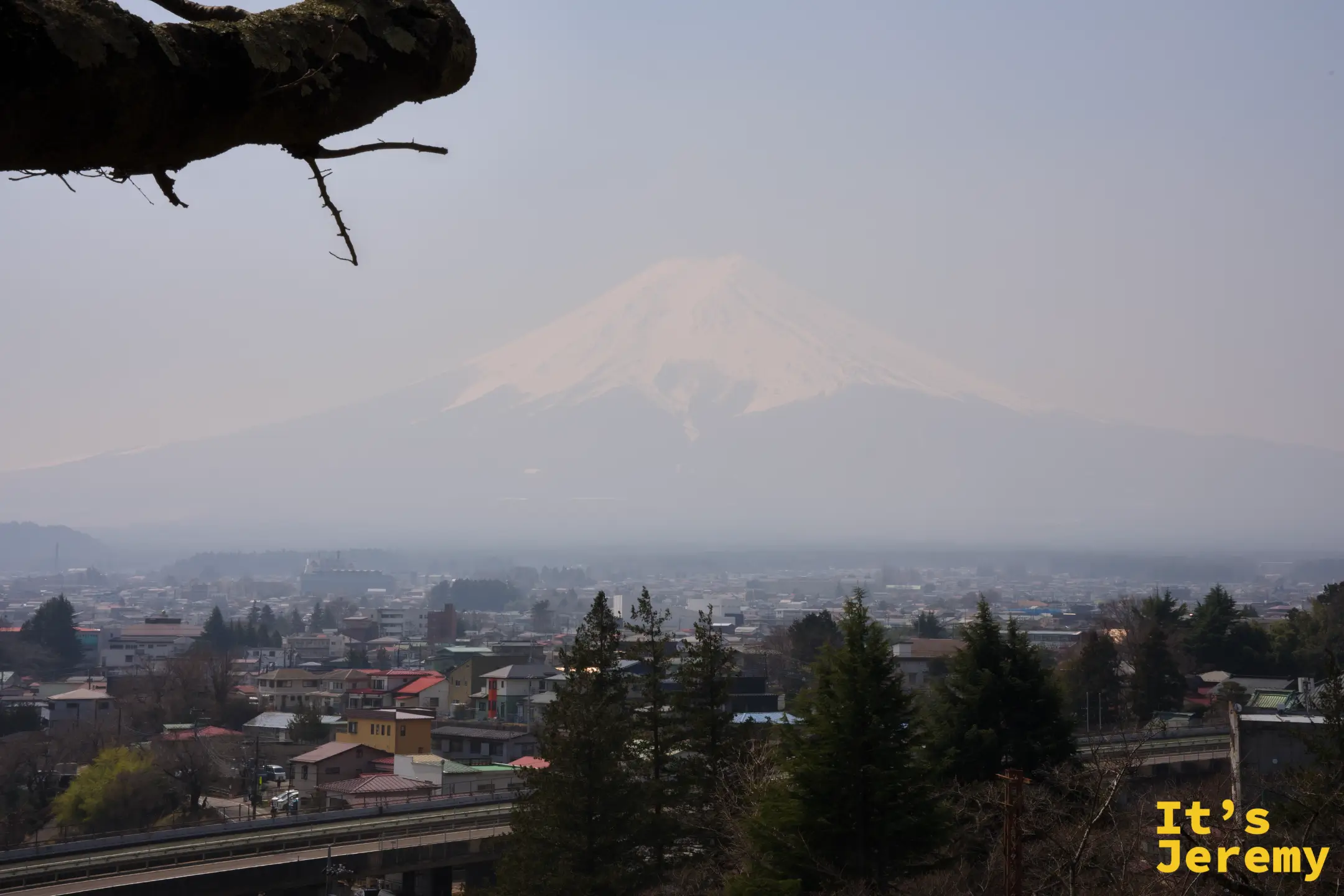 從神社中段平台俯瞰下吉田市區，房屋與橋梁清晰，富士山在沙塵裡化成淡色輪廓
