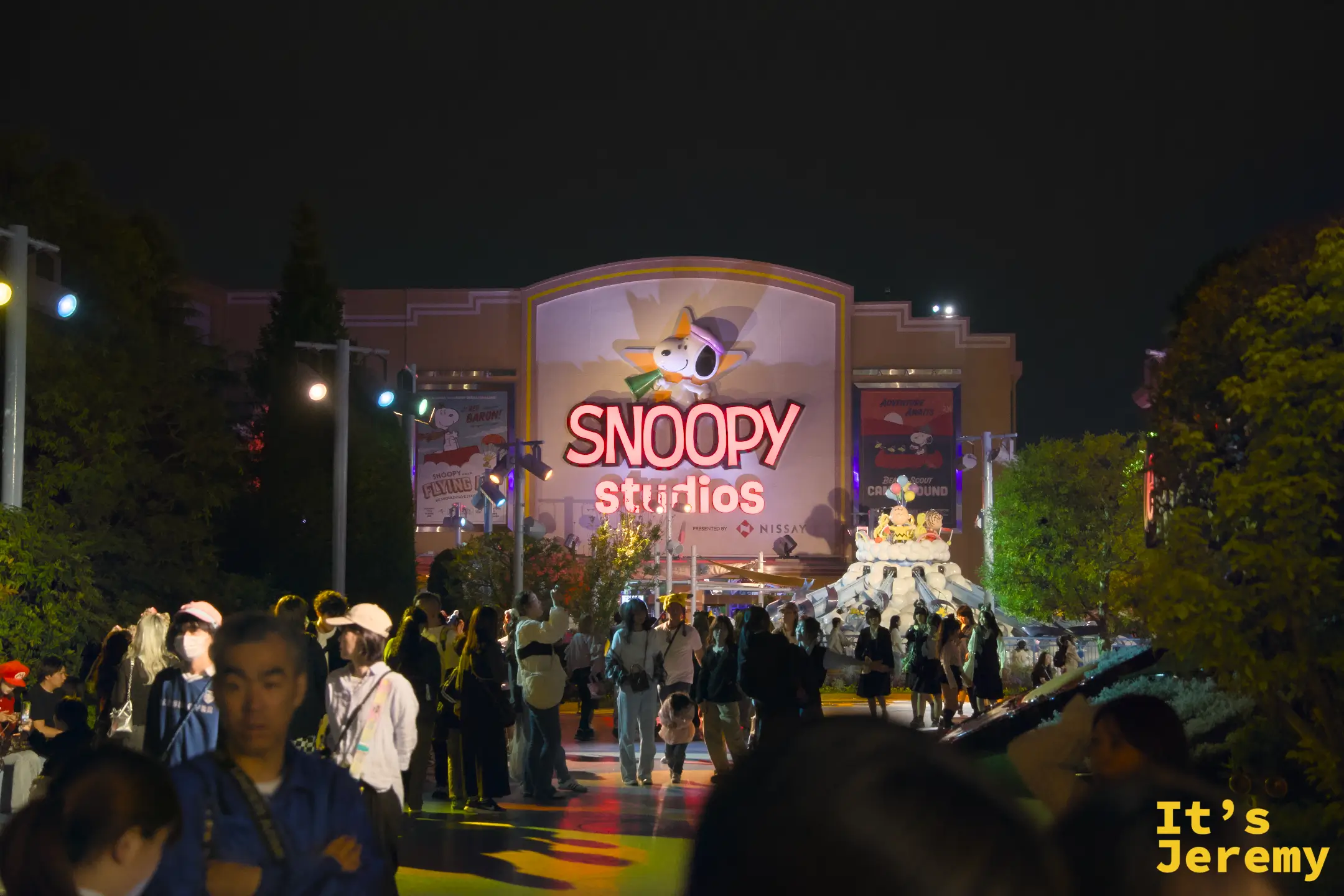 Snoopy Studios 夜景，霓虹燈管組成的 Snoopy 圖案在建築正面閃爍，旁邊有 25 週年白色蛋糕裝置，入口廣場人潮絡繹不絕