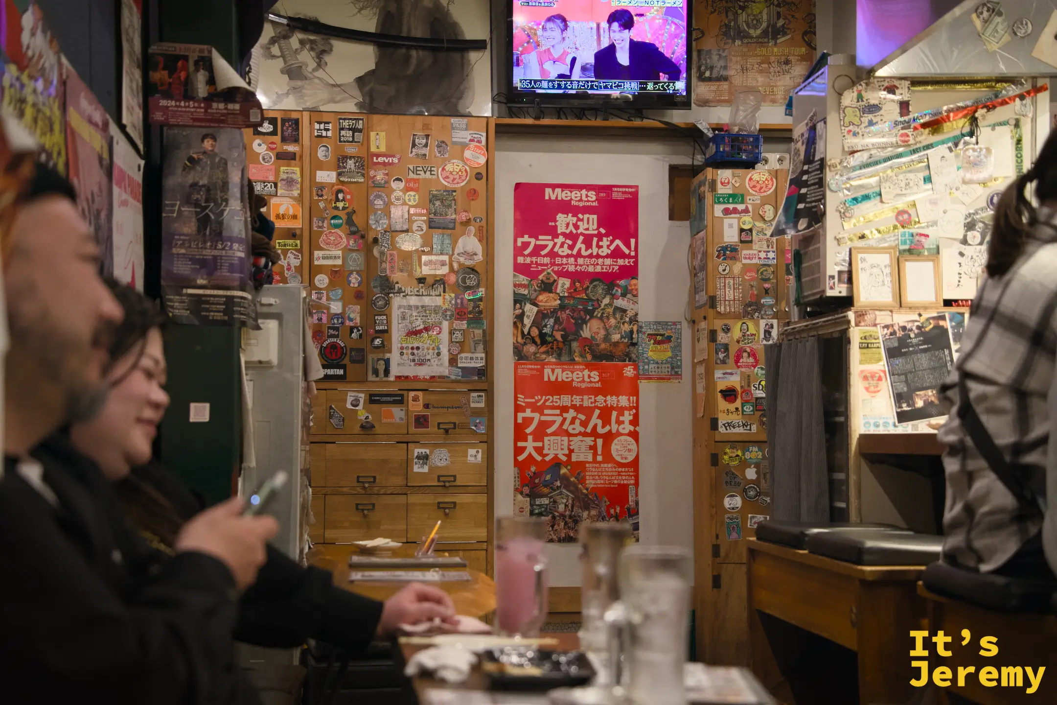 客人在吧台前聊天大笑，背後貼著「歡迎，ウラなんばへ！」Meets 雜誌海報，電視在角落播放