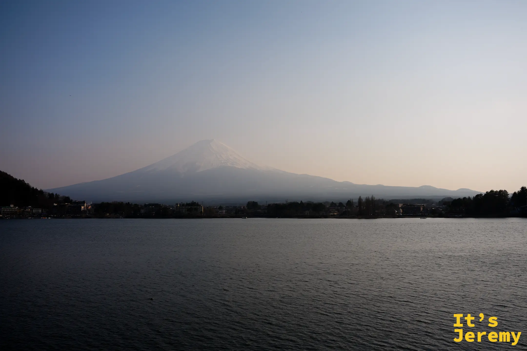 傍晚的河口湖，富士山輪廓開始從沙塵中浮現，湖面深藍、山腰仍帶霾