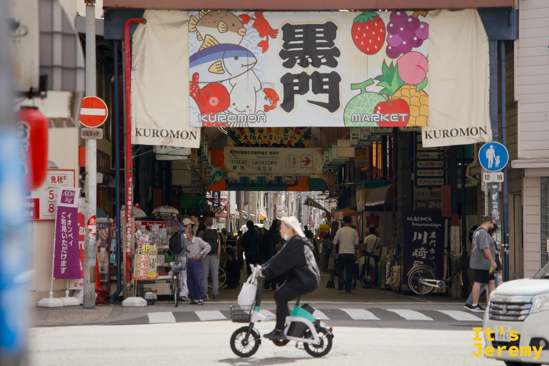 市場入口近拍，招牌下騎腳踏車的行人與周邊店家