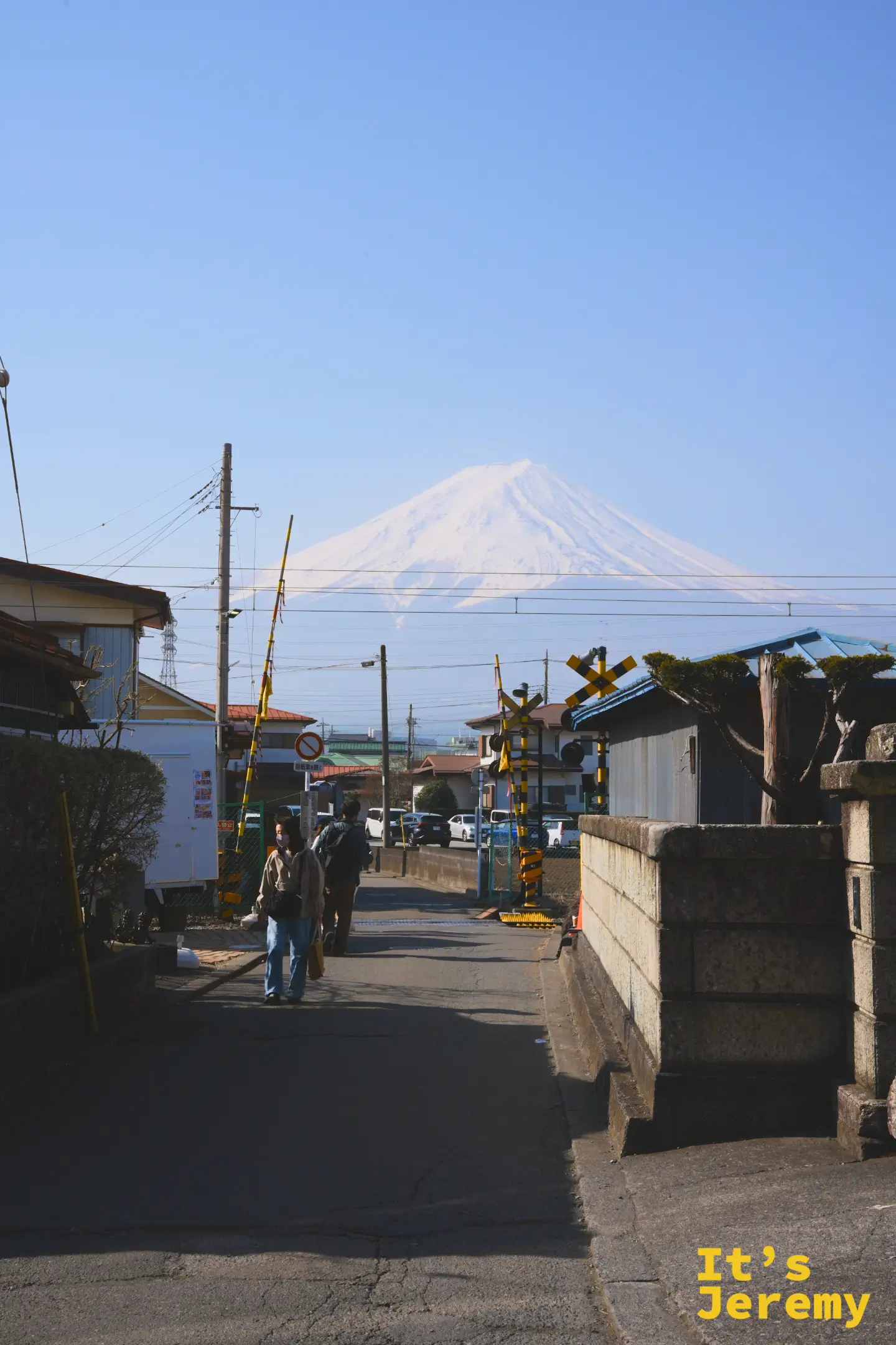 平交道欄杆旁，電線杆與住宅屋頂之間，富士山戴著雪帽矗立在清晨的藍天下