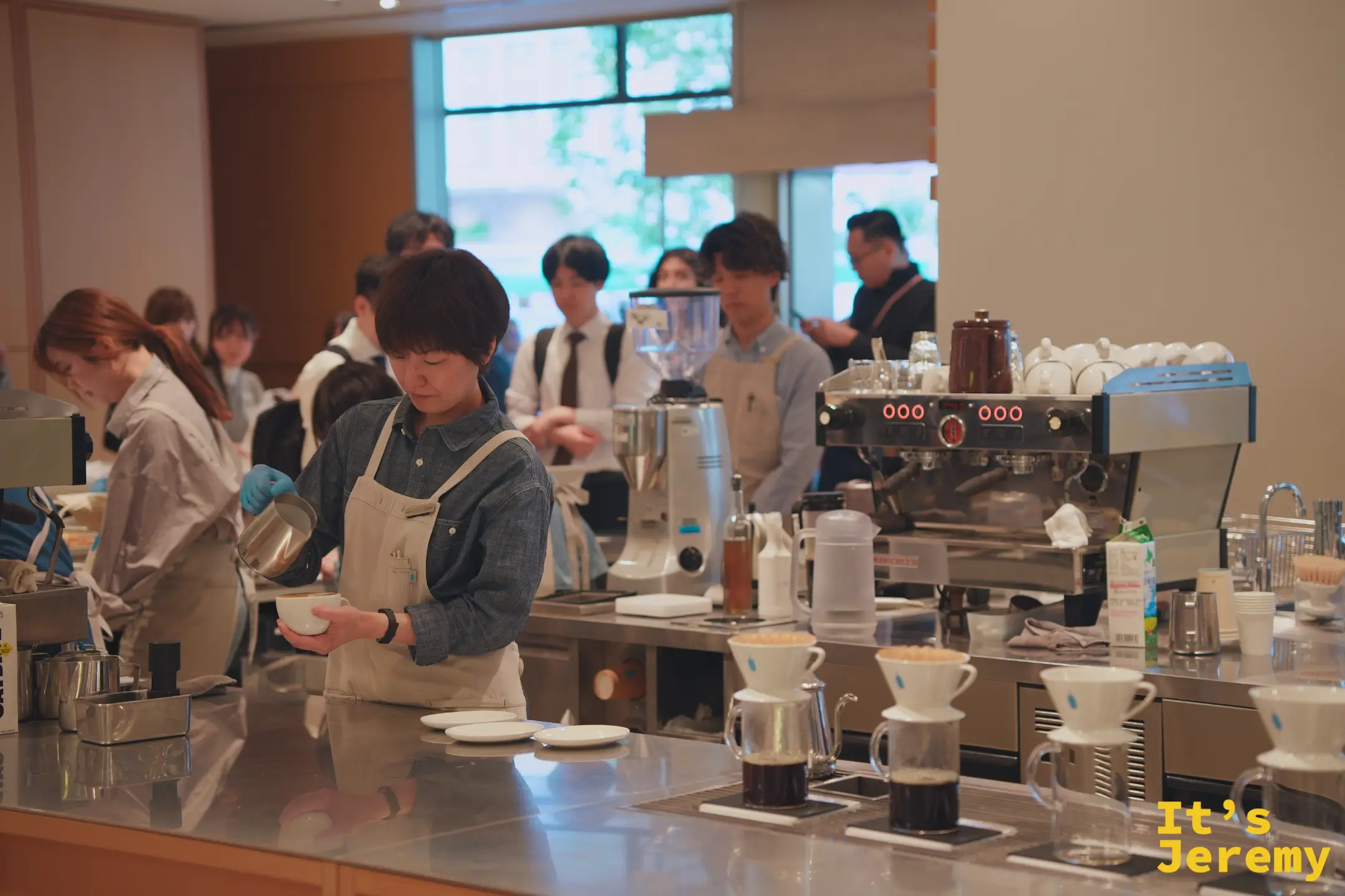 Blue Bottle 新宿店吧台後方，數名身穿牛仔圍裙的咖啡師同步操作，手沖架上排著整列 V60 濾杯與玻璃分享壺