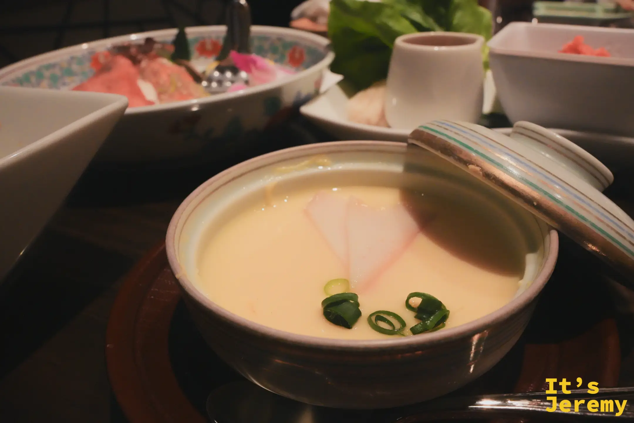 茶碗蒸裝在有蓋陶碗內，蛋黃色蛋液上有蔥段與粉紅魚板