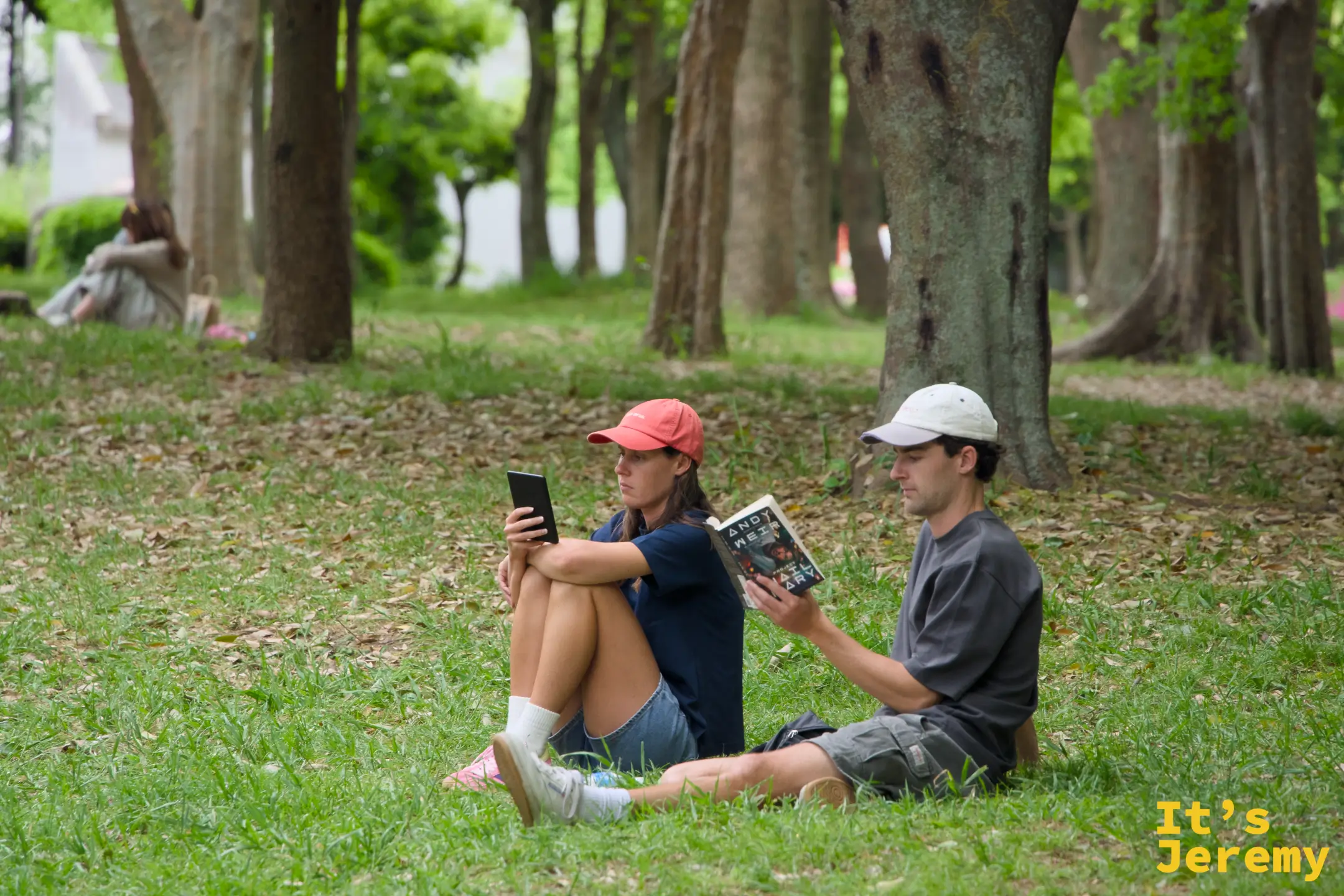 兩人坐在公園草地的樹蔭下，一人看手機、一人看書