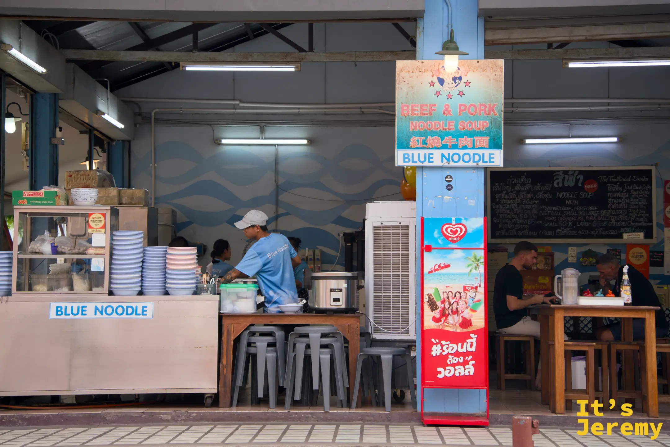 店面外觀，備餐台左側寫著 Blue Noodle，右側客人正在用餐