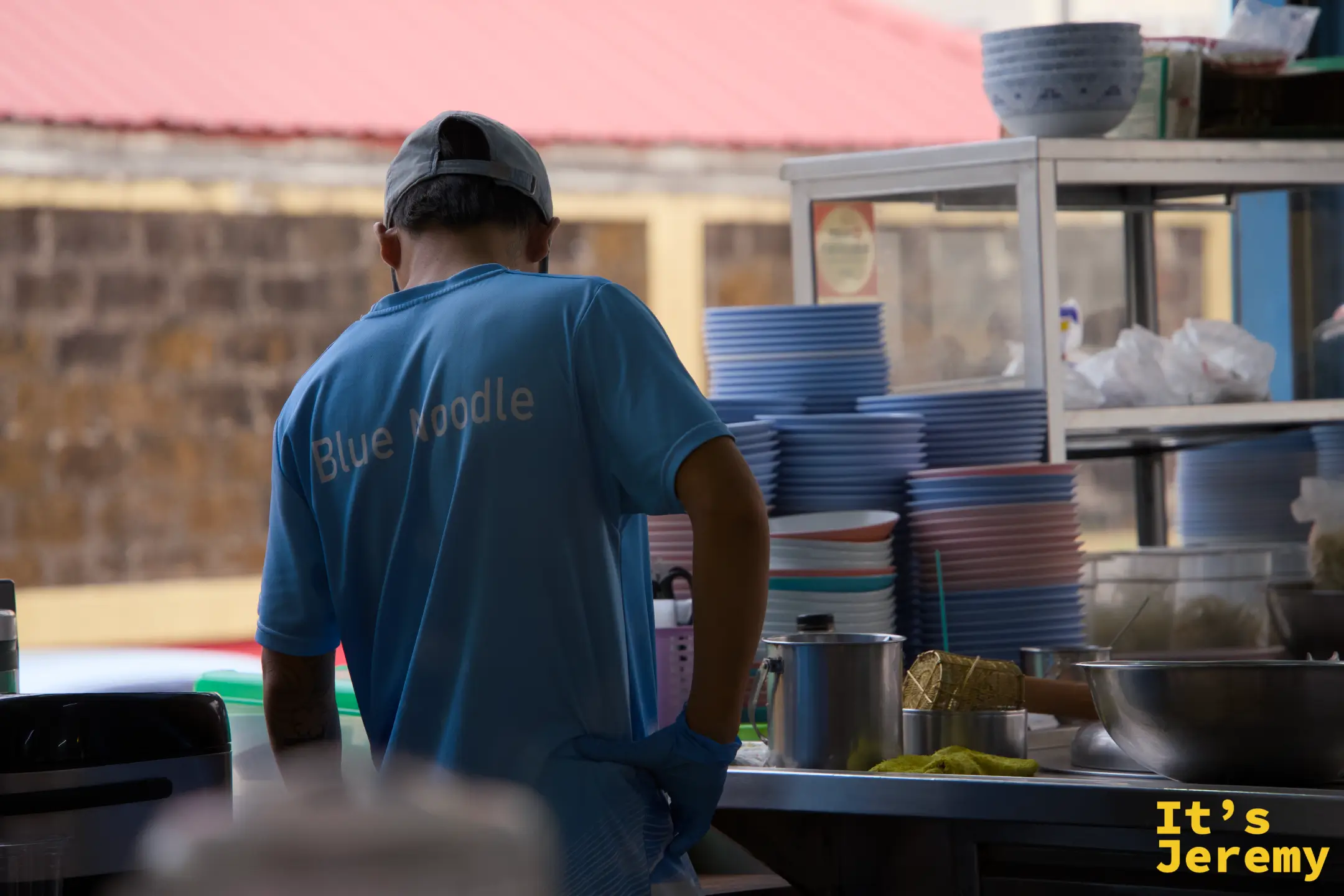 穿 Blue Noodle 制服的男店員在廚房備餐，背後是整齊堆疊的藍色碗盤