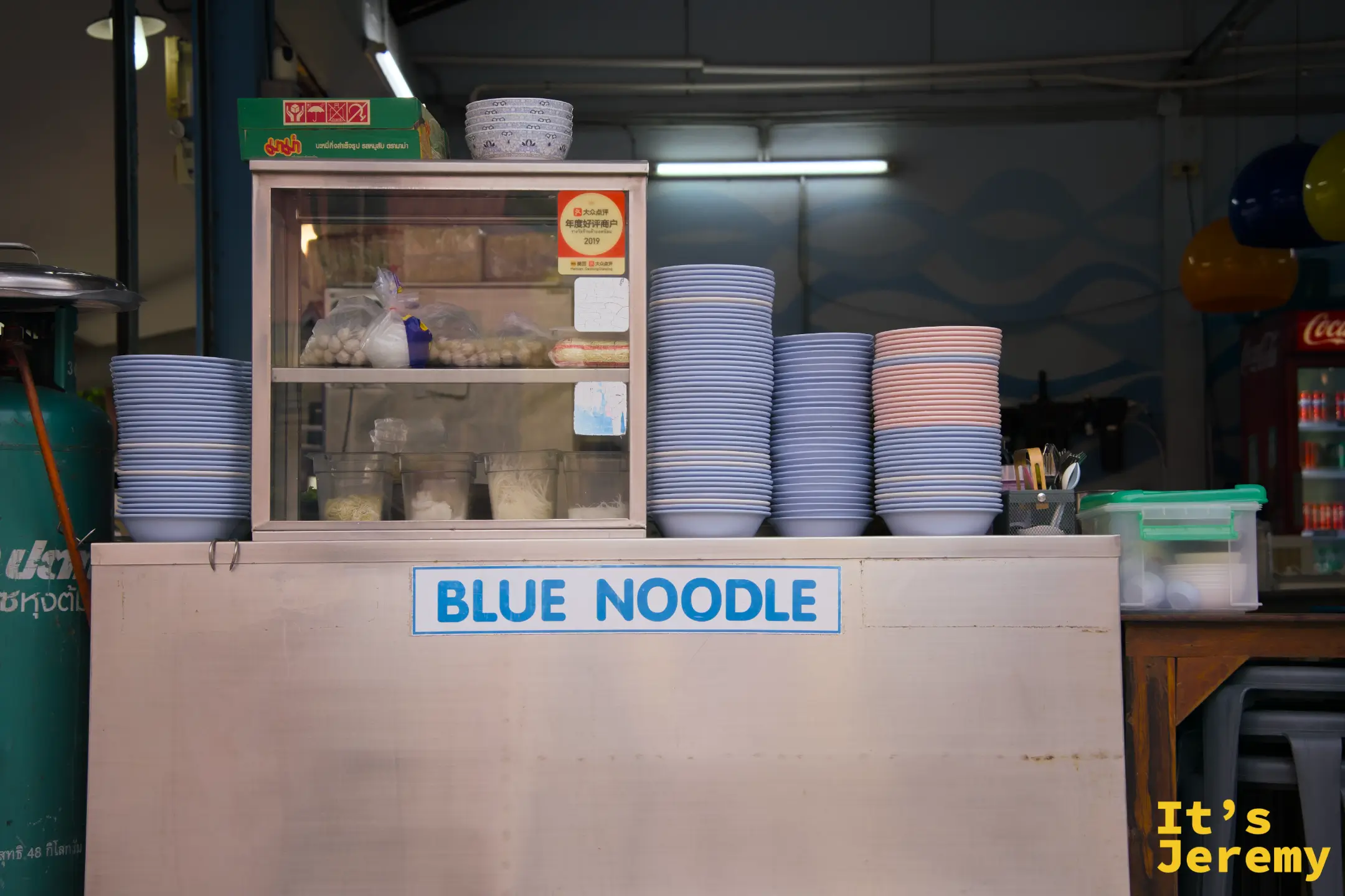 Blue Noodle 備餐台，台面寫著品牌名稱，上方整齊堆放大量藍色與粉色碗盤