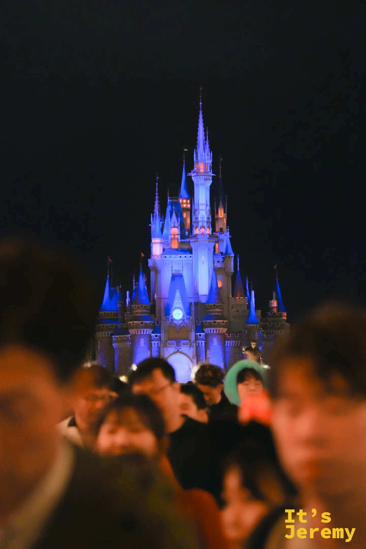 夜間點燈的城堡，前景是戴著米奇耳朵的人群剪影