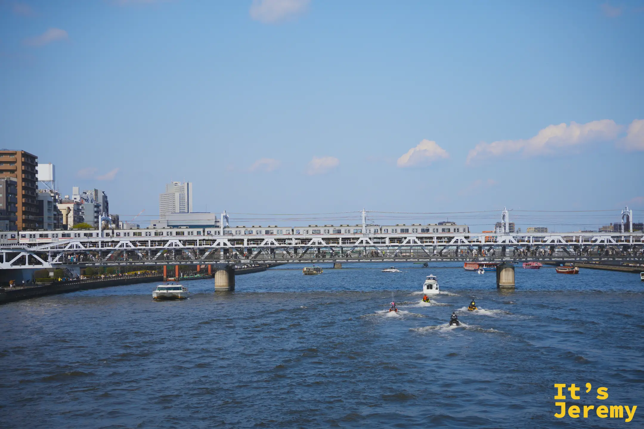 隅田川寬闊的河面，鐵路橋上有列車正在通過，河上有水上摩托車和遊船穿梭