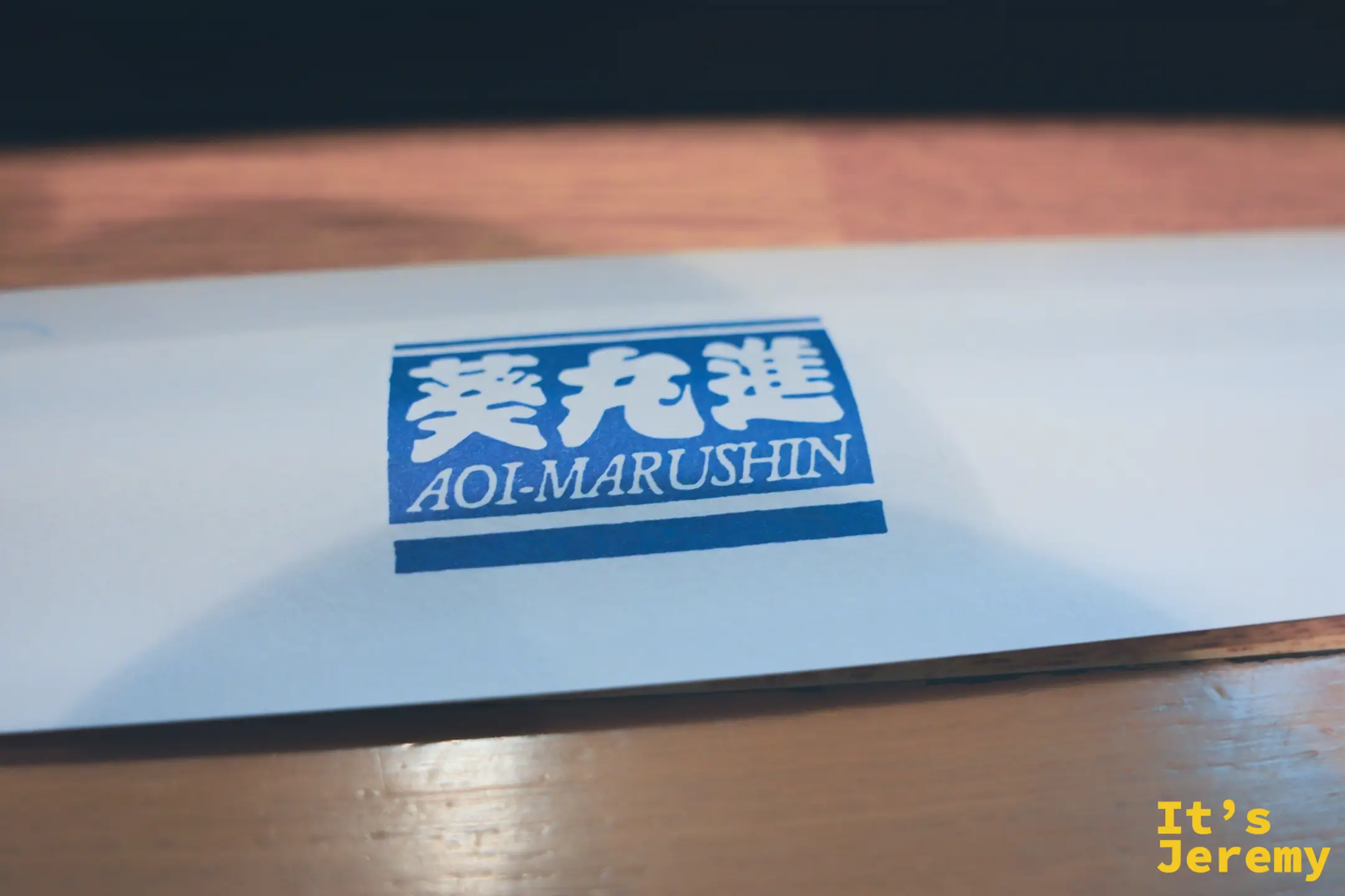葵丸進的白底藍字品牌紙袋平放在木質桌面上，印著葵丸進漢字與「AOI-MARUSHIN」英文字樣