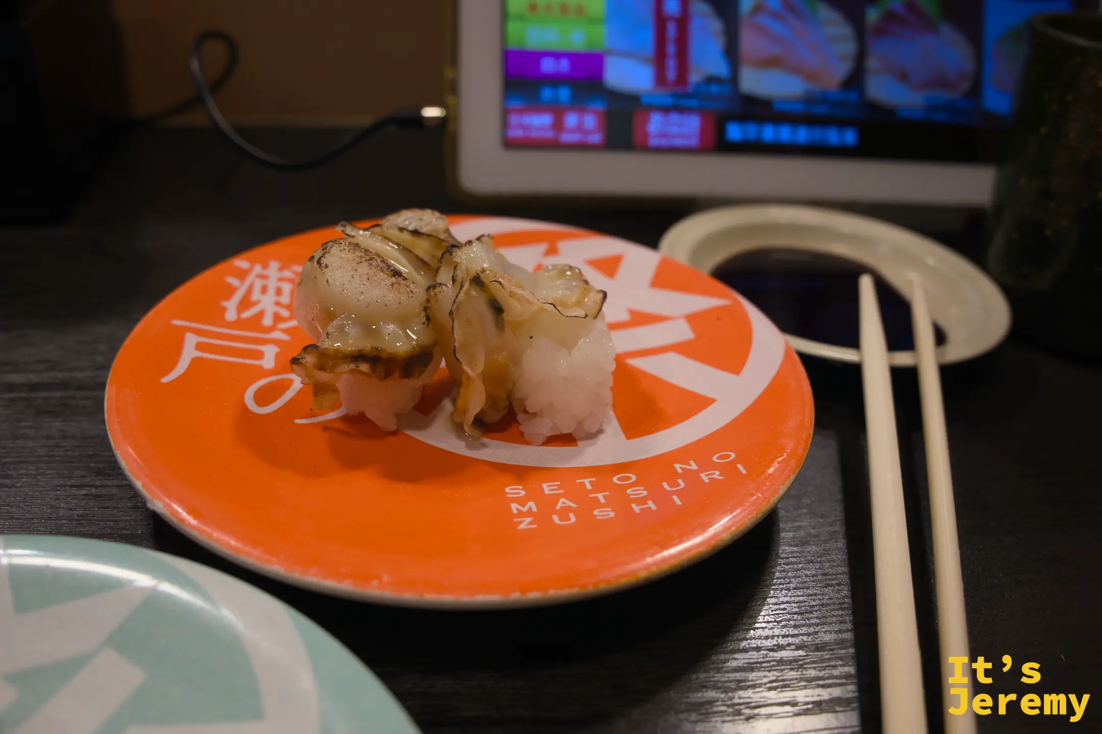橘紅色盤上的炙燒帆立貝握壽司，貝肉表面有焦紋，盤緣印「SETO NO MATSURI ZUSHI」