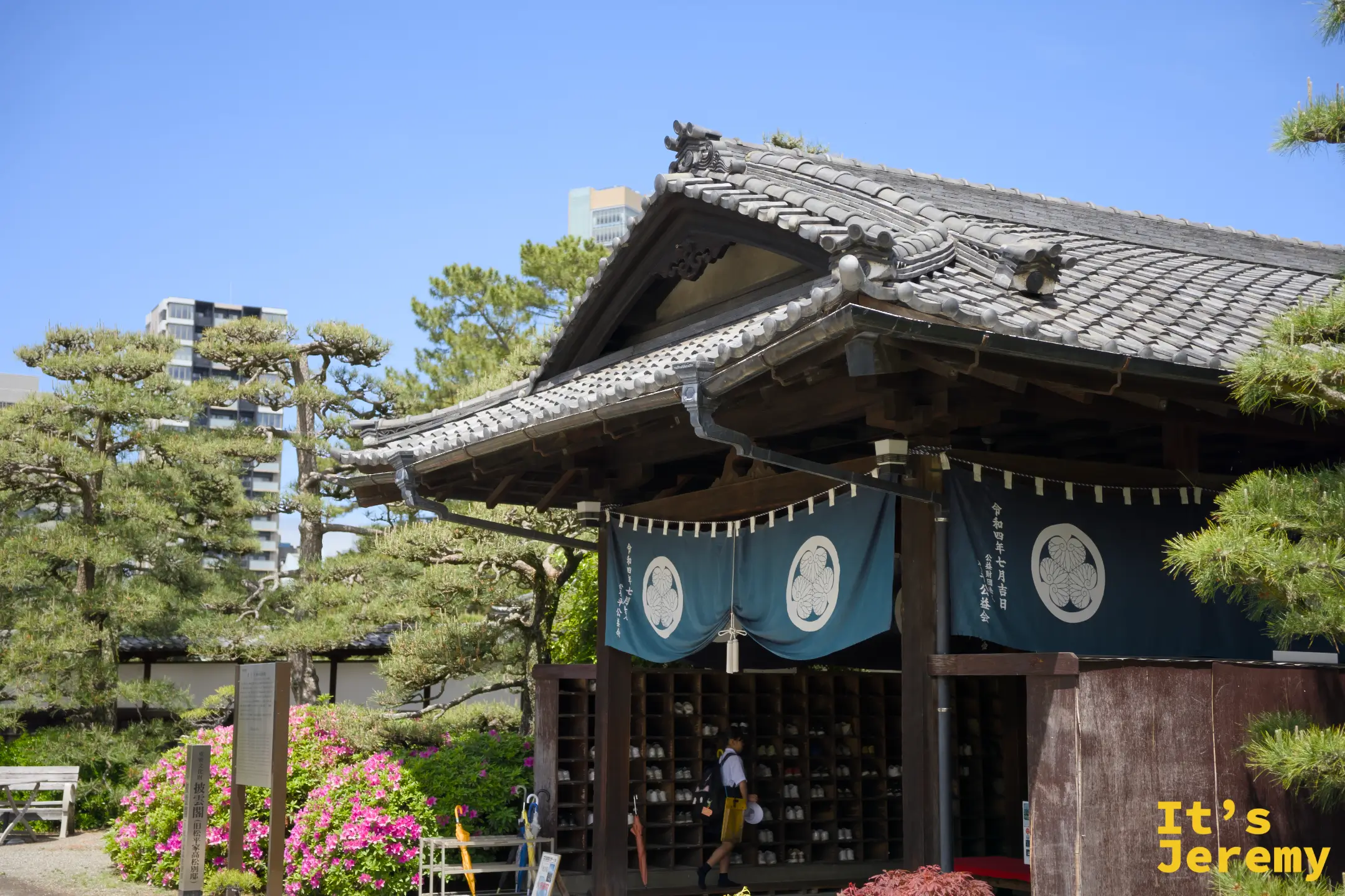 晴天下的披雲閣入口全景，杜鵑花正盛開，對稱的黑松守著兩側，藍色松平家紋幕布高掛大門