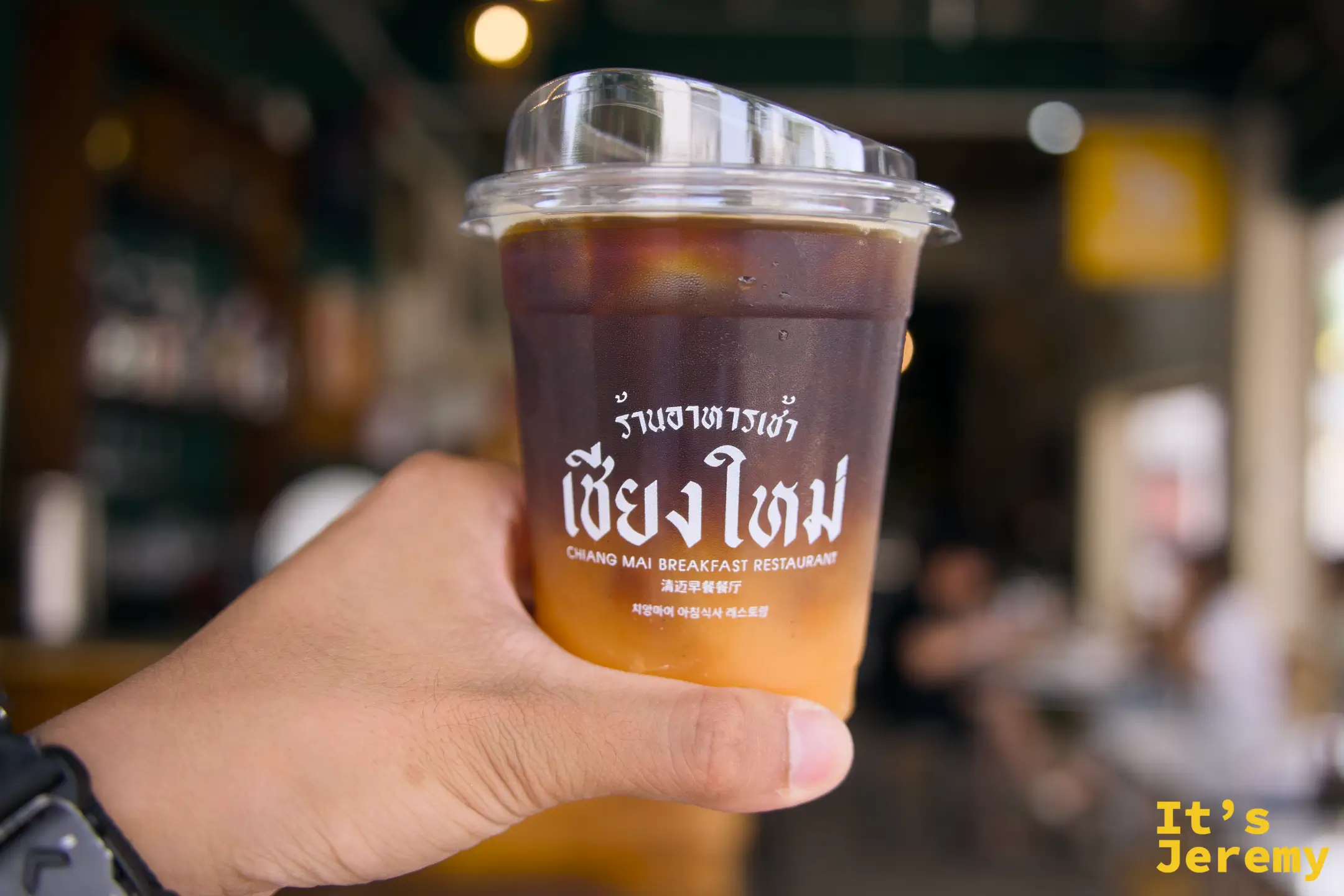 手持印有清邁早餐餐廳 Logo 的 Orange Americano 透明杯，背景店內空間