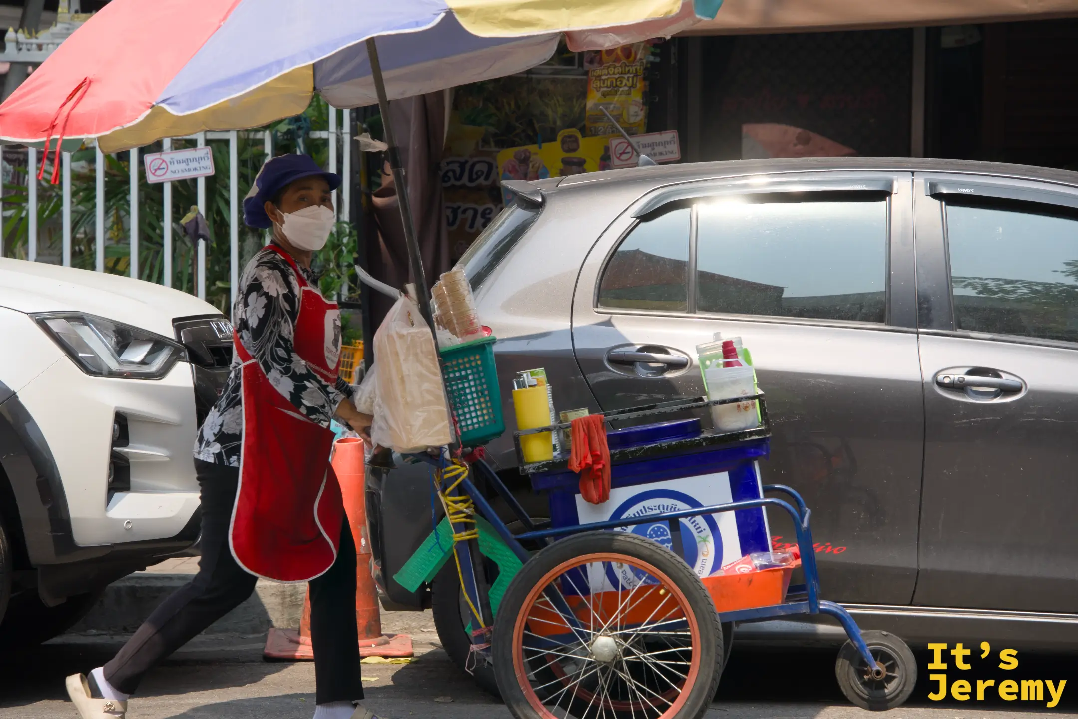 穿紅色圍裙的街頭小販推著三輪餐車，撐一把大傘，走在路上攬客