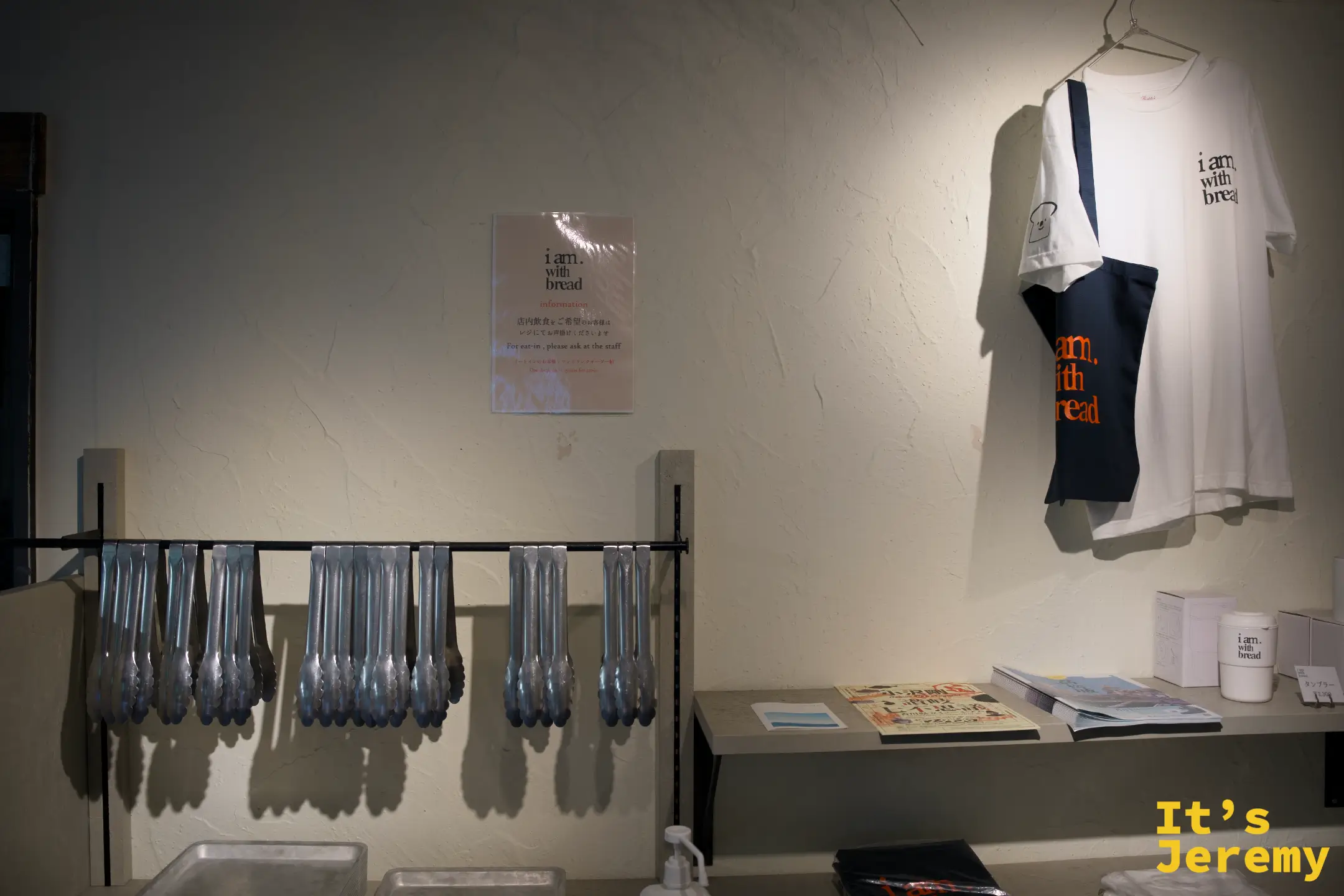 店內牆邊掛著印有品牌 logo 的白色 T 恤與托特包，下方鐵架掛著一整排餐具