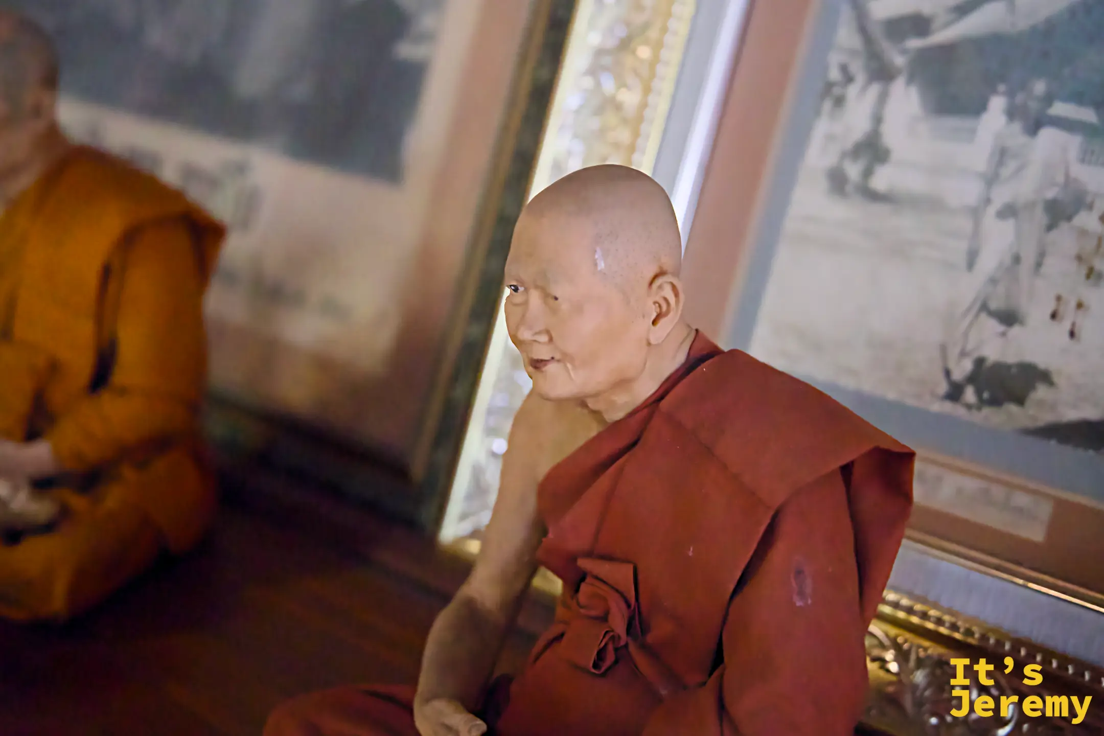 高僧蠟像，面部細節高度逼真，穿著橘色僧衣，室內光線昏暗，氛圍肅穆