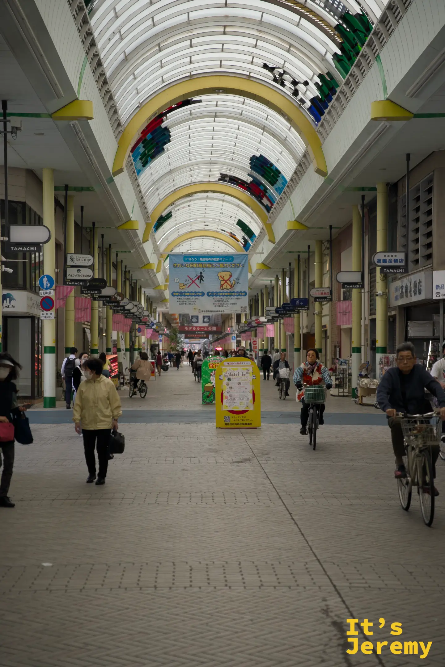 高松田町商店街（TAMACHI）內部，拱形頂棚覆蓋的長廊兩側商家林立，市民騎著自行車穿梭其中