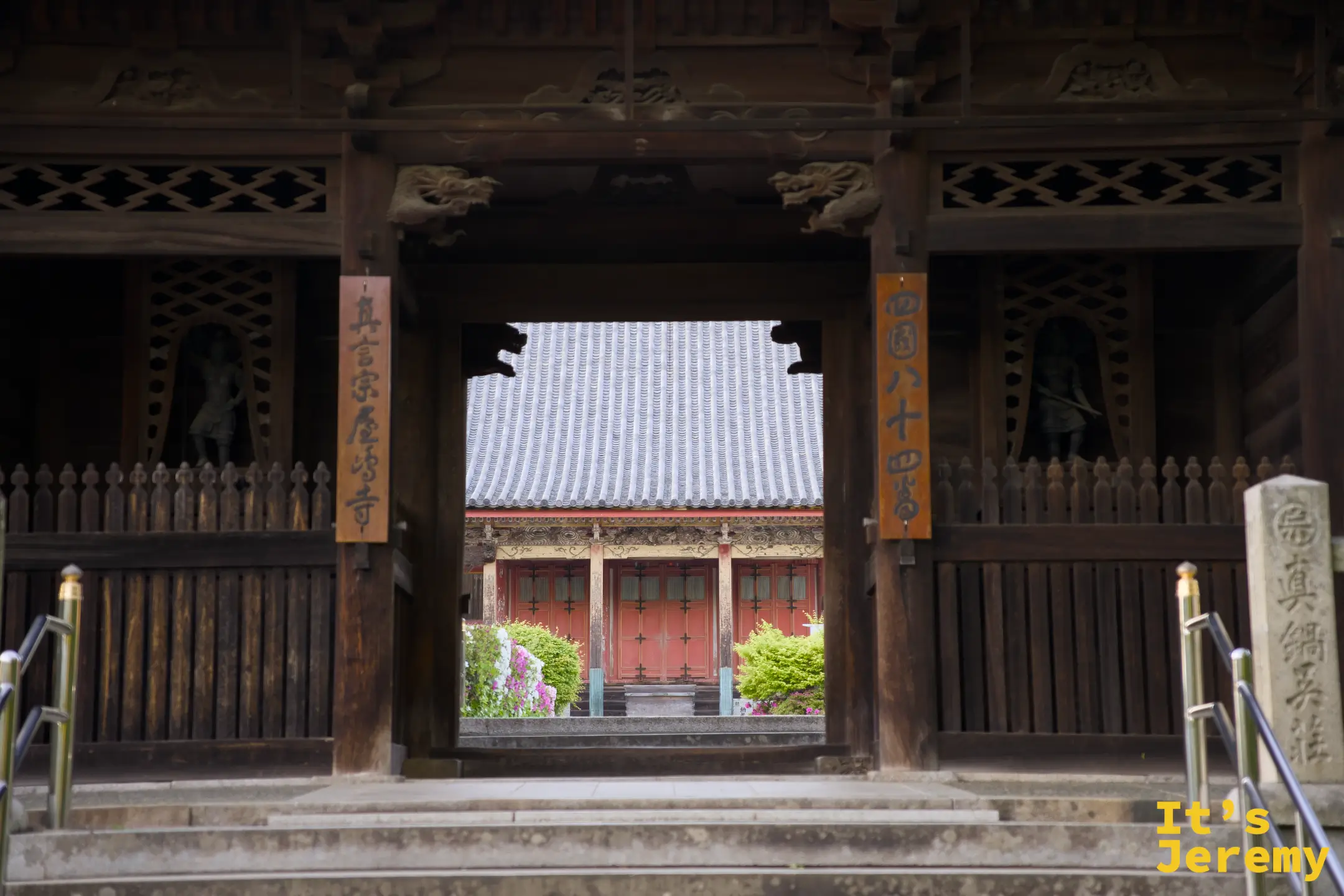 兩位遊客沿石板小徑走向屋島寺山門，綠葉在光線下泛著金黃，一幅安靜的午後參道