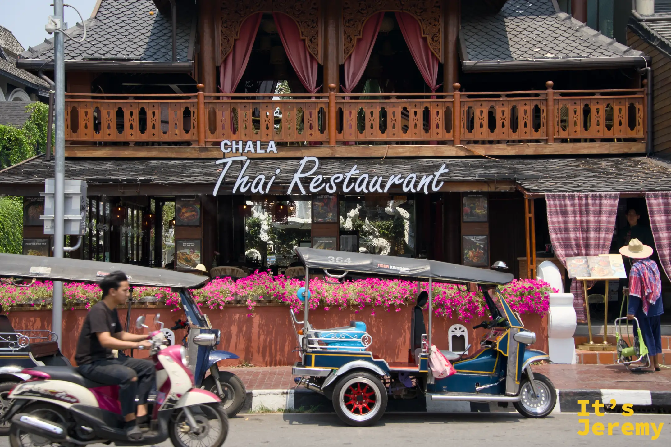 CHALA Thai Restaurant 木造傳統泰式建築外觀，橘紅花叢前停著一輛裝飾嘟嘟車，機車穿越畫面