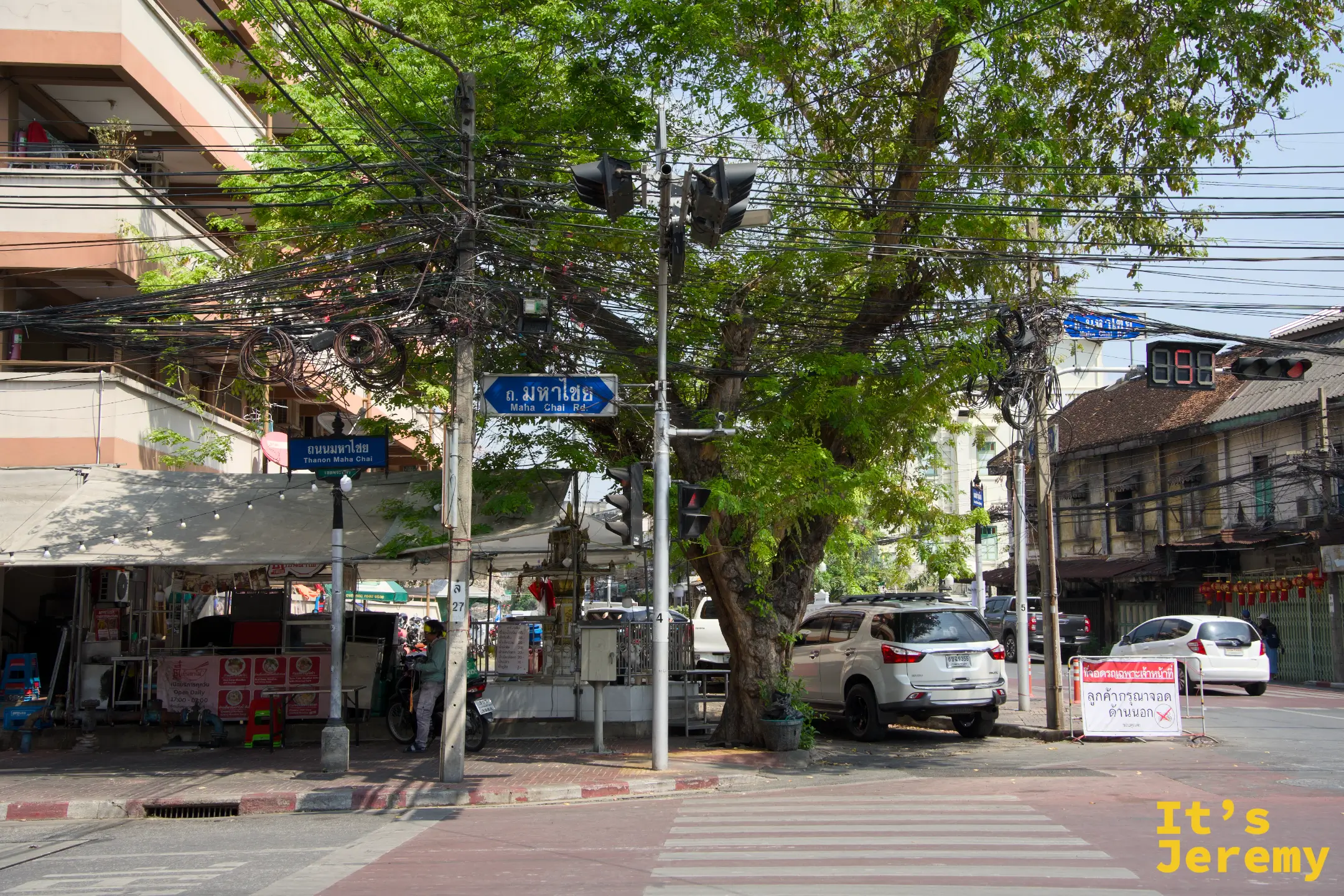 另一個角度的 Mahachai 路口街景，藍底路牌與綠樹下的老城區午後