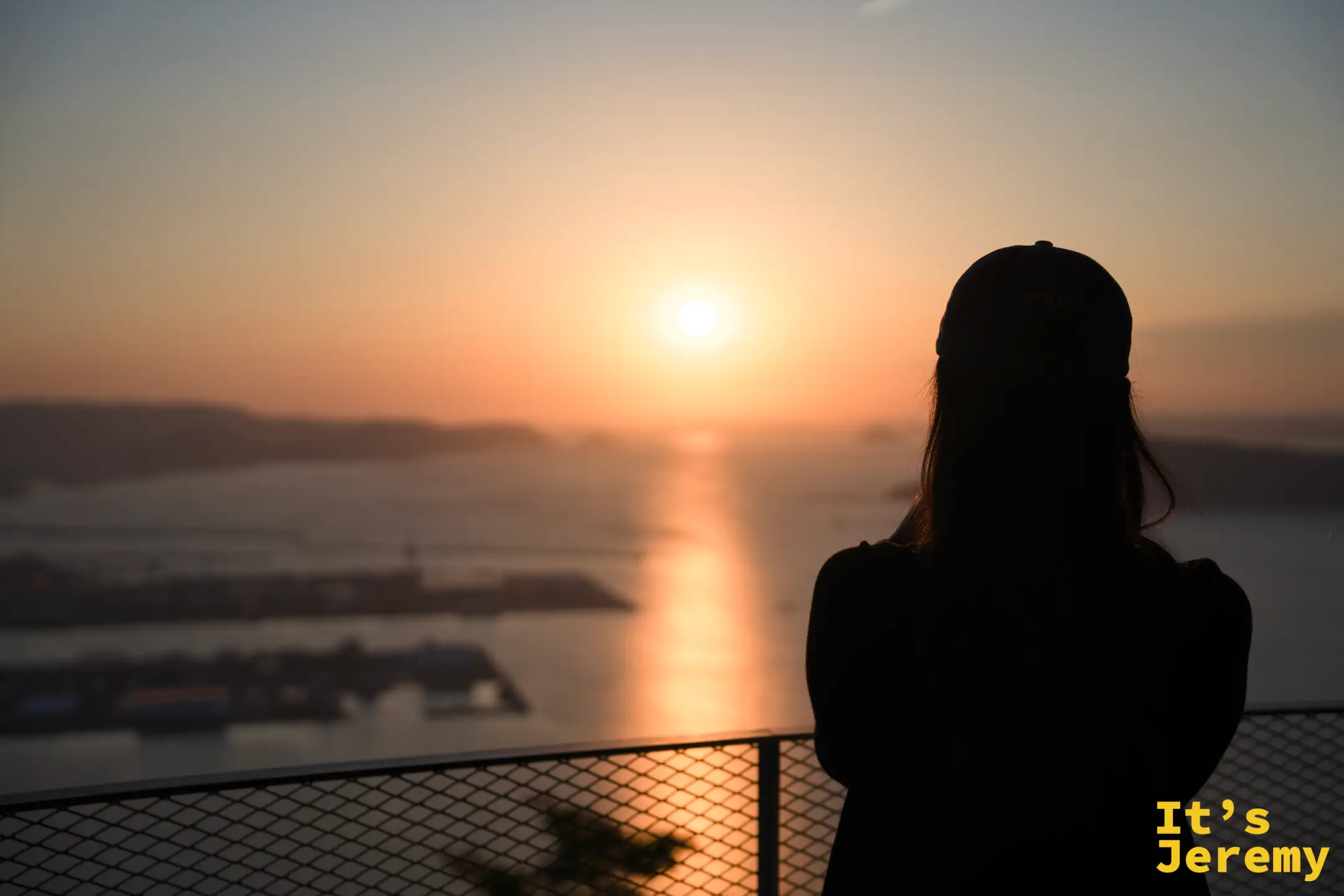 展望台欄杆邊，一位戴帽女性背對鏡頭凝視日落，瀨戶內海島嶼在她身後的橙色光裡展開