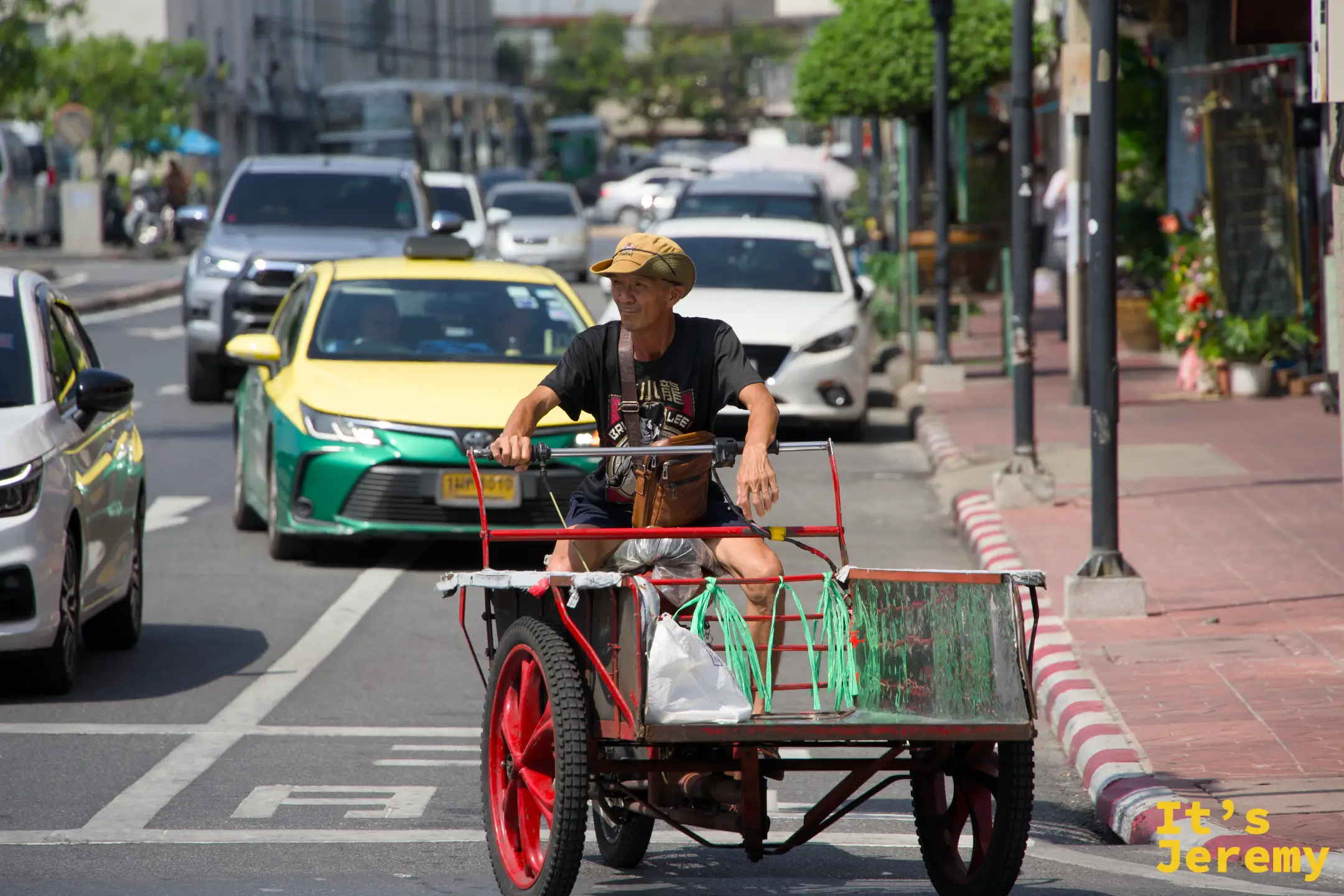 一位老先生戴著迷彩帽推著木板車過馬路，車上載著綠色塑膠袋——老城區最日常的搬運畫面