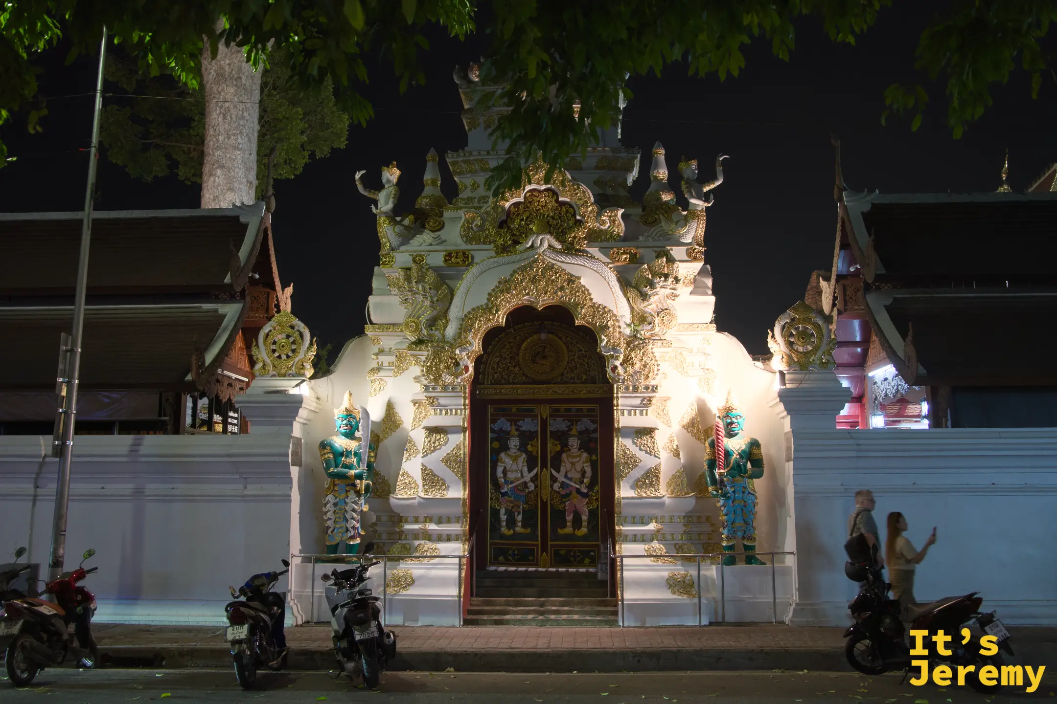 夜深後山門緊閉,燈光打亮白色金飾與兩旁夜叉,路人走過,廟宇歸於安靜