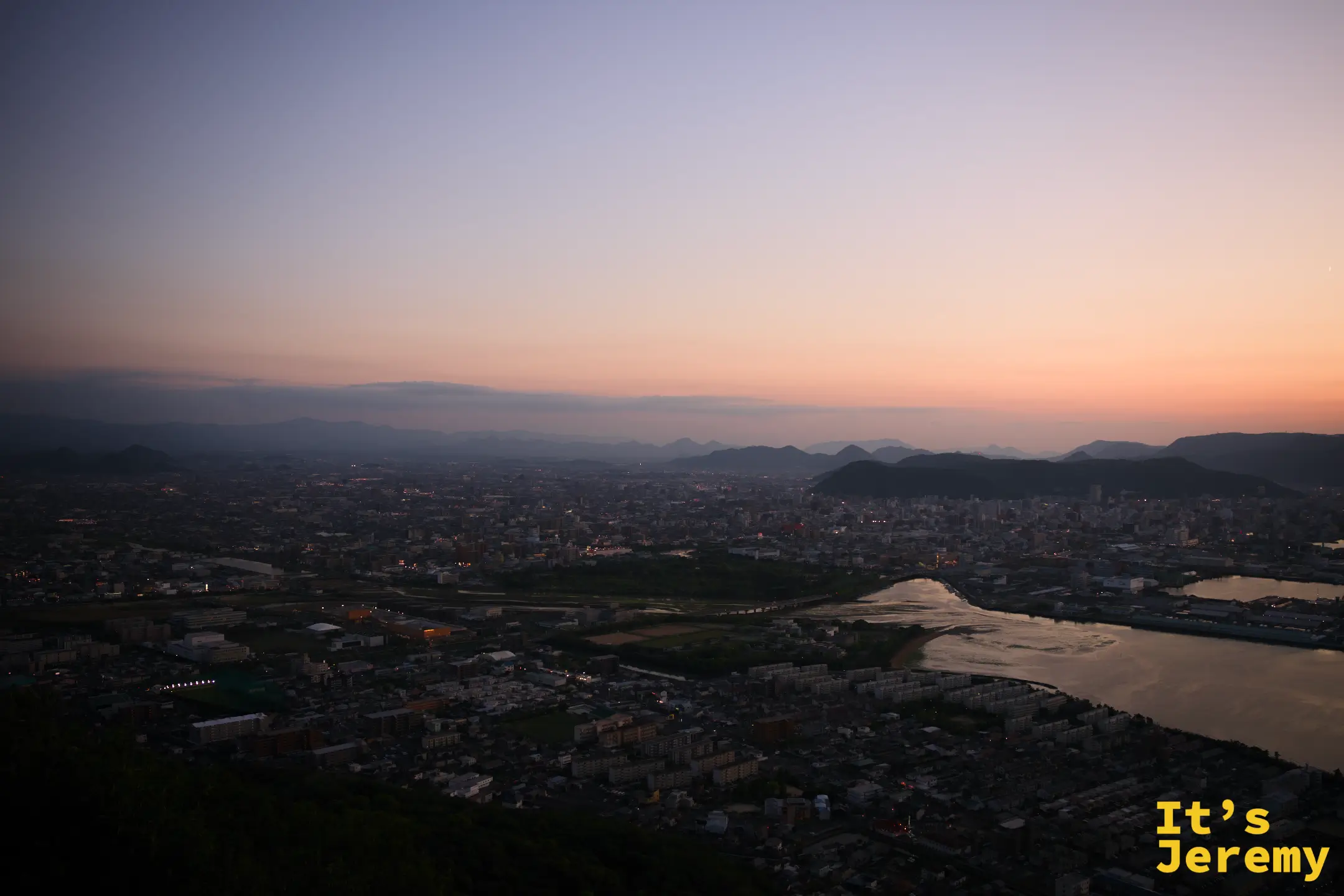 高松市區廣角夜景，蓬萊川蜿蜒貫穿，遠山疊層，城市燈光在藍調中星星點點亮起