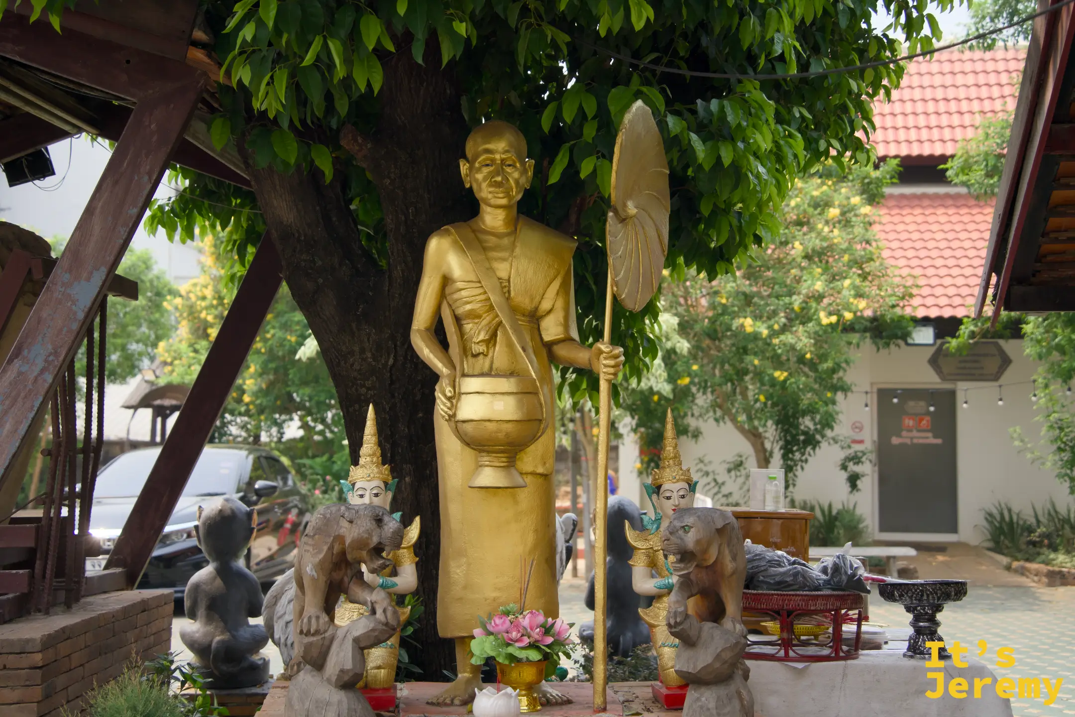 金色站立高僧雕像，左持托缽右持傘，身旁有數尊小型侍者石雕圍繞，前方有鮮花供品