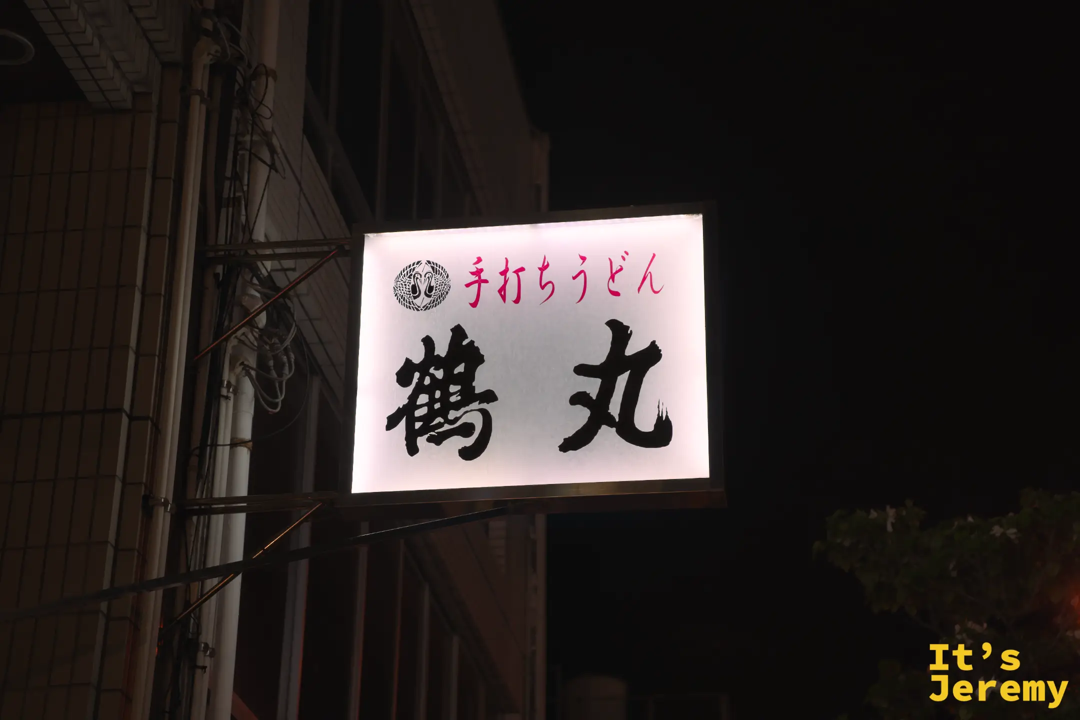 夜晚的鶴丸招牌燈箱，白底黑字「鶴丸」，上方紅字「手打ちうどん」