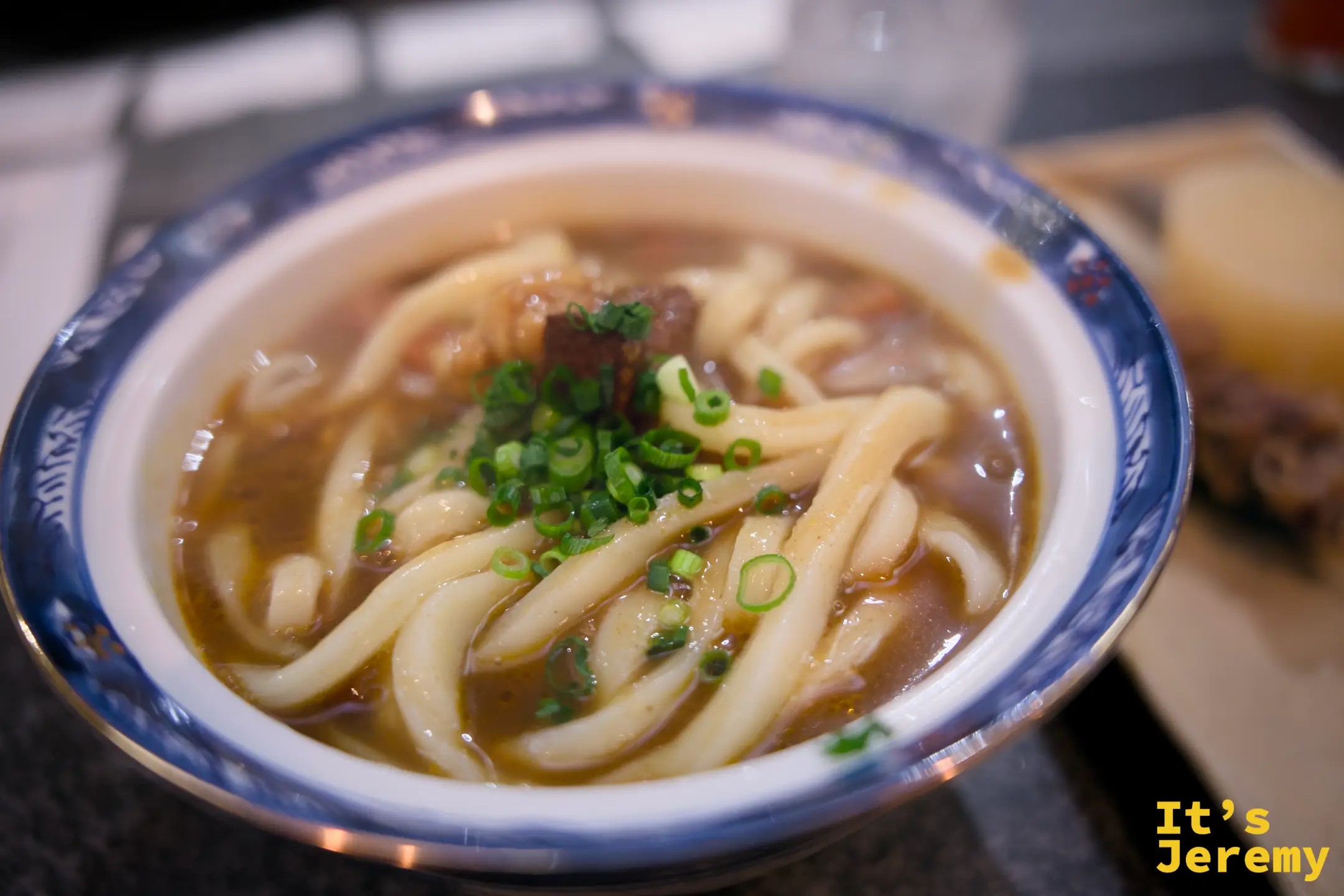 咖哩烏龍麵特寫，深棕色咖哩高湯，粗韌麵體，上頭撒一把碧綠蔥花