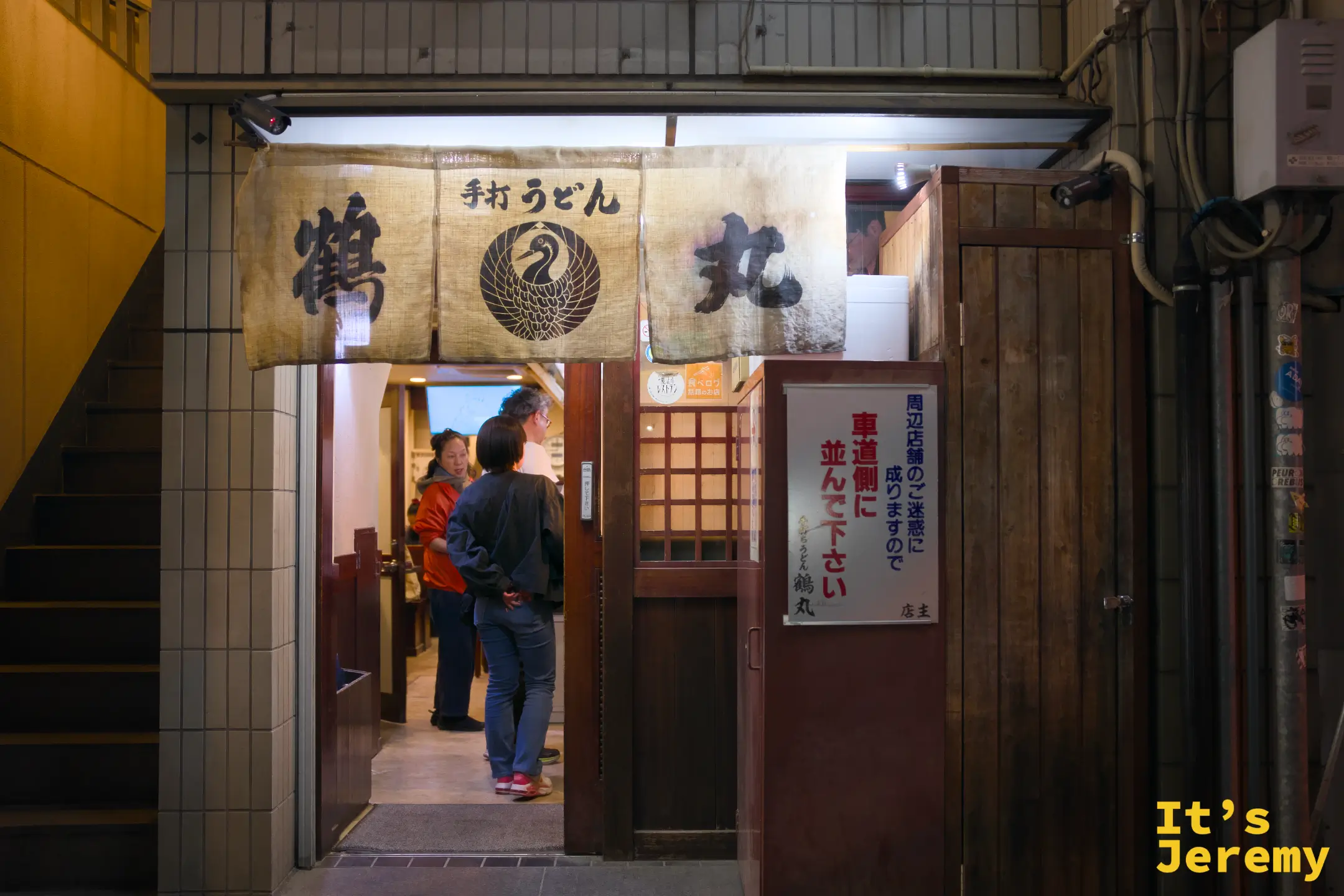 夜晚街邊的烏龍麵店入口，布製暖簾印著「手打うどん 鶴丸」，店內燈光溫暖
