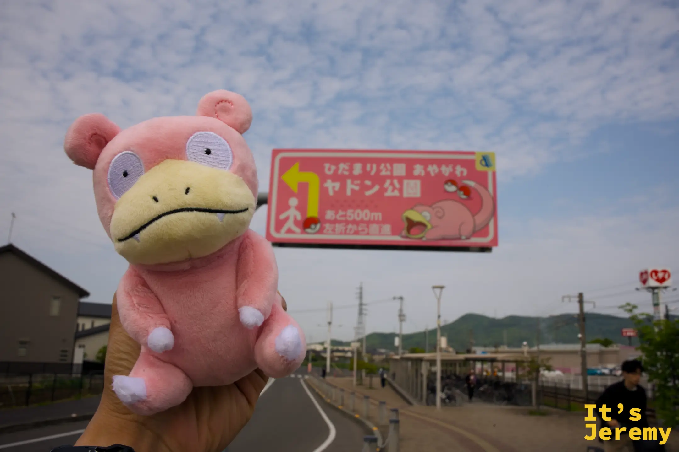 手持呆呆獸玩偶在指路牌旁合影，後方是綾川町的住宅區與遠山