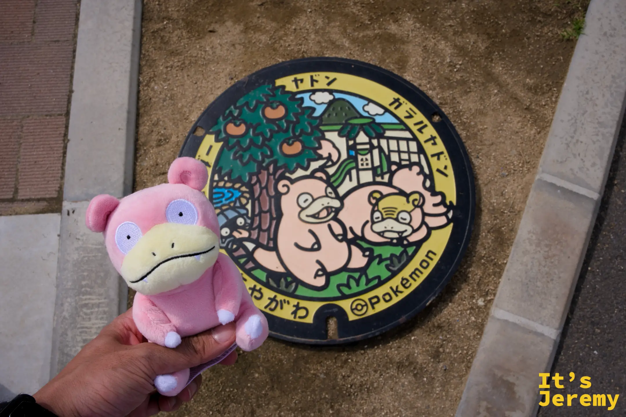 黃邊人孔蓋前，手持呆呆獸布偶對齊畫面，蓋面繪有呆呆獸與伽勒爾呆呆獸，背景為橘子樹與古民家