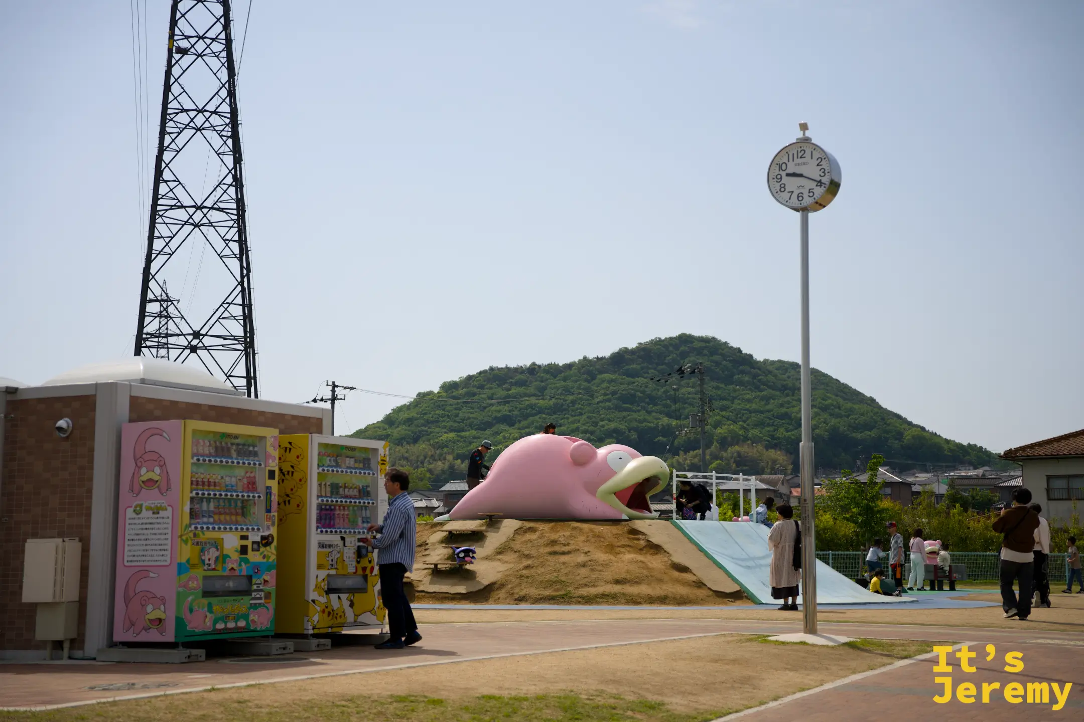 呆呆獸公園全景，左邊是兩台寶可夢販賣機與圓形時鐘柱，中央是巨型趴臥呆呆獸，遠方是丘陵